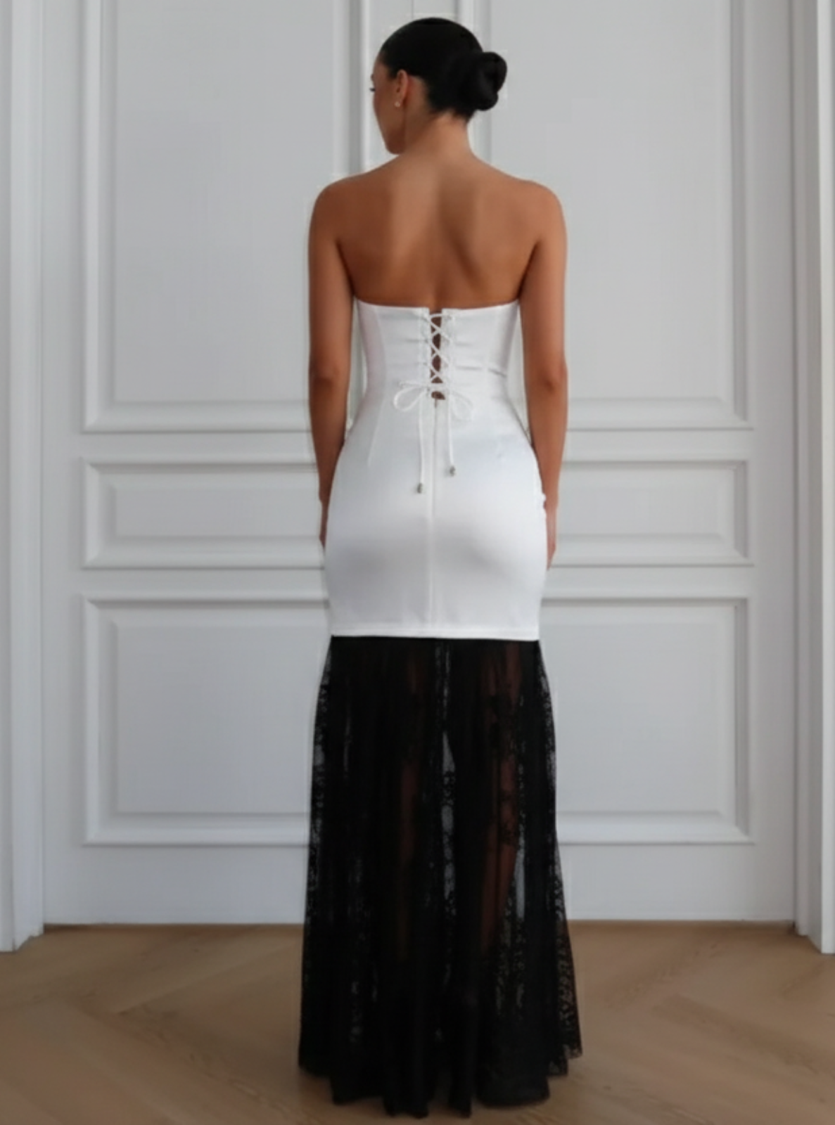 Woman wearing a white strapless top and black sheer skirt against a white paneled wall.