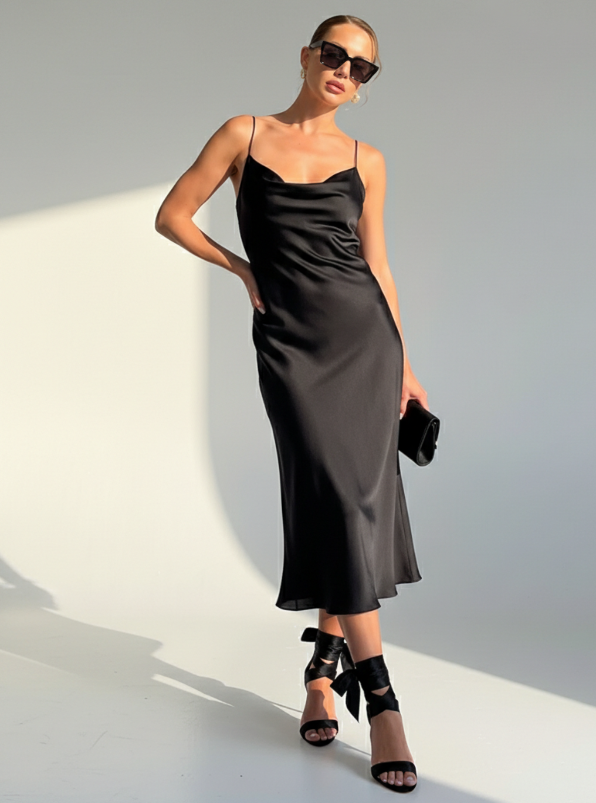 Woman wearing a black slip dress with sunglasses and high heels on a white background