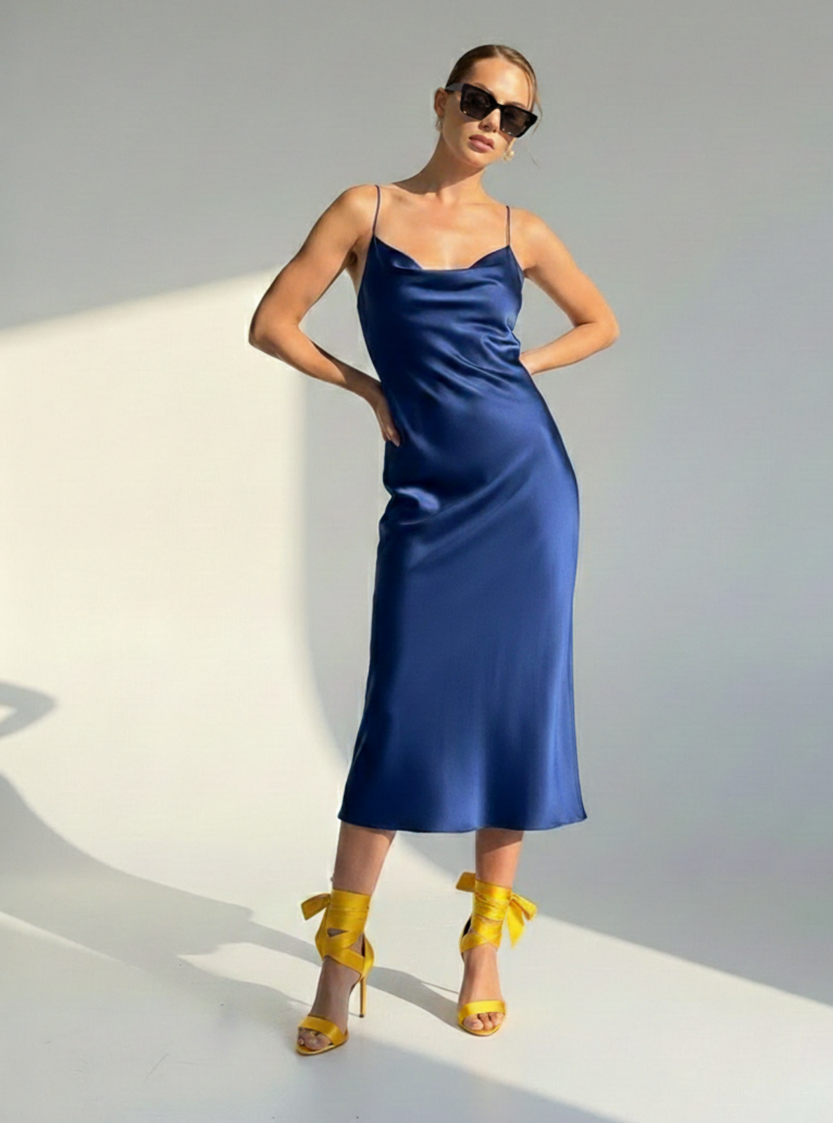 Woman wearing a blue satin dress with yellow shoes and sunglasses on a light background