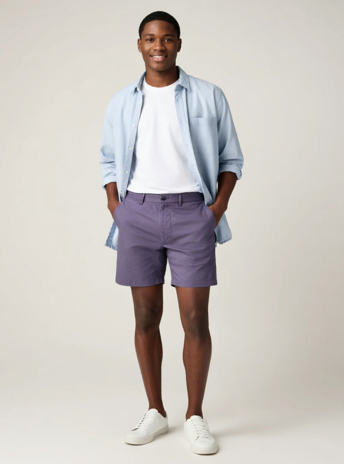 Man wearing a light blue shirt, white t-shirt, purple shorts, and white sneakers on a plain background