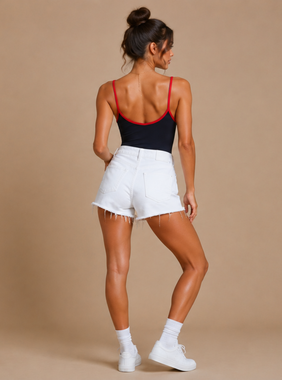 A woman is shown from behind wearing a sleeveless black tank top with red trim, white denim shorts, white socks, and white sneakers, standing against a solid beige studio backdrop.