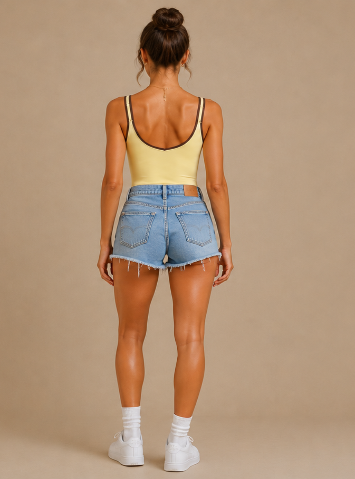A woman shown from the back wearing a pale yellow tank top with black trim and light blue denim shorts, standing against a solid beige studio background.