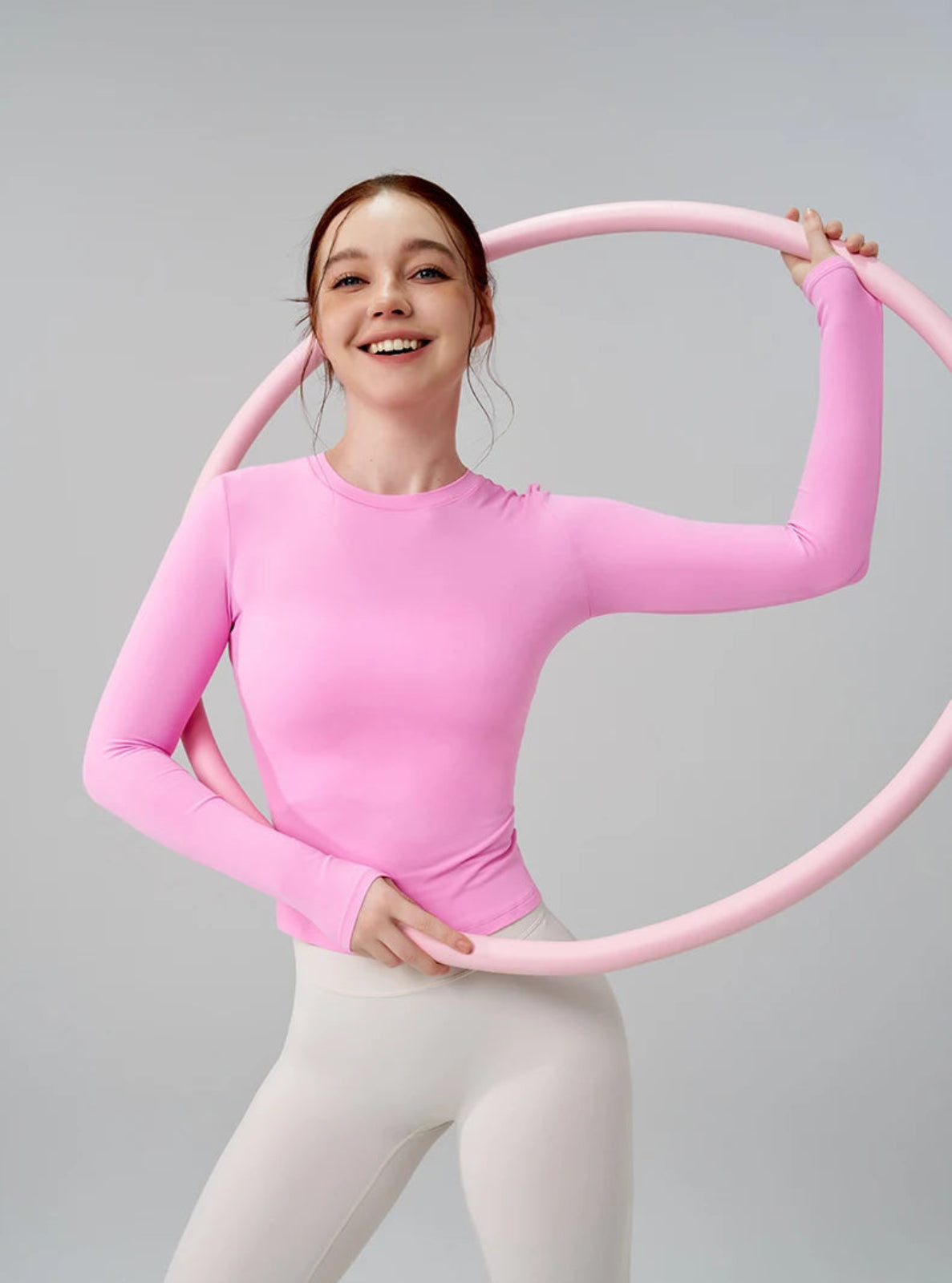 Person wearing a pink long-sleeve top and white pants holding a pink hula hoop against a gray background
