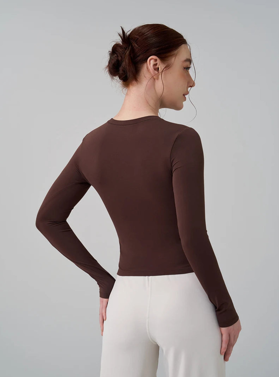 Woman wearing a brown long-sleeve top and white pants against a gray background