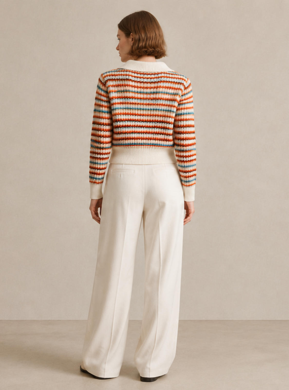 A woman is shown from behind wearing a cropped, long-sleeve knit sweater with horizontal stripes in orange, red, and blue, paired with high-waisted, wide-leg white trousers, set against a neutral beige studio backdrop.