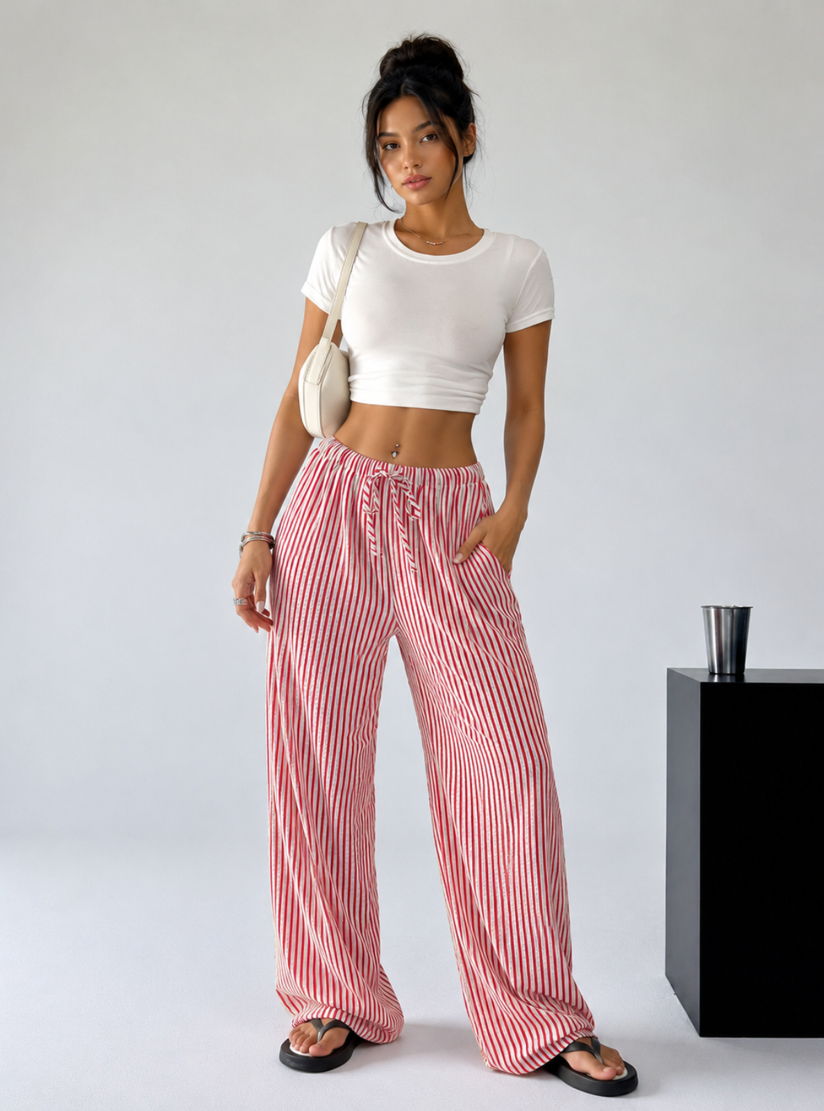 A woman modeling a white crop top and pink and white vertically striped wide-leg pants, standing in a studio setting next to a black pedestal.