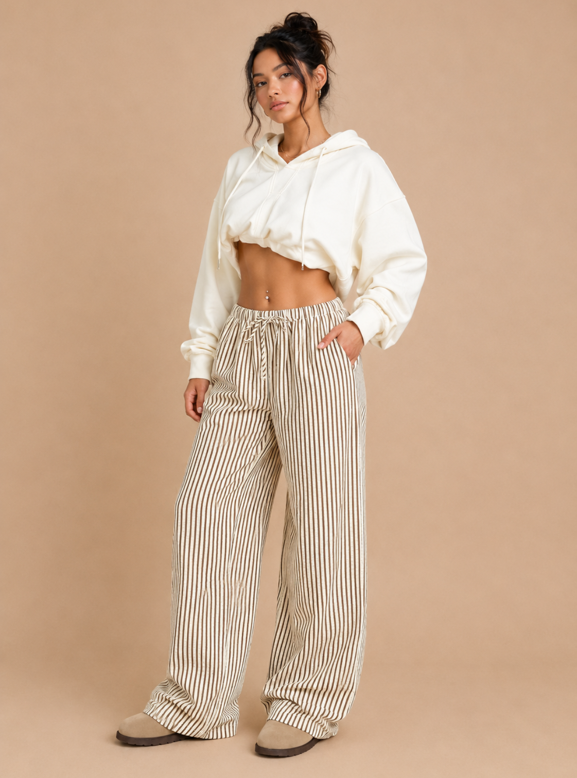 A woman modeling a white cropped hoodie and beige-and-white vertically striped wide-leg pants against a solid beige studio background.