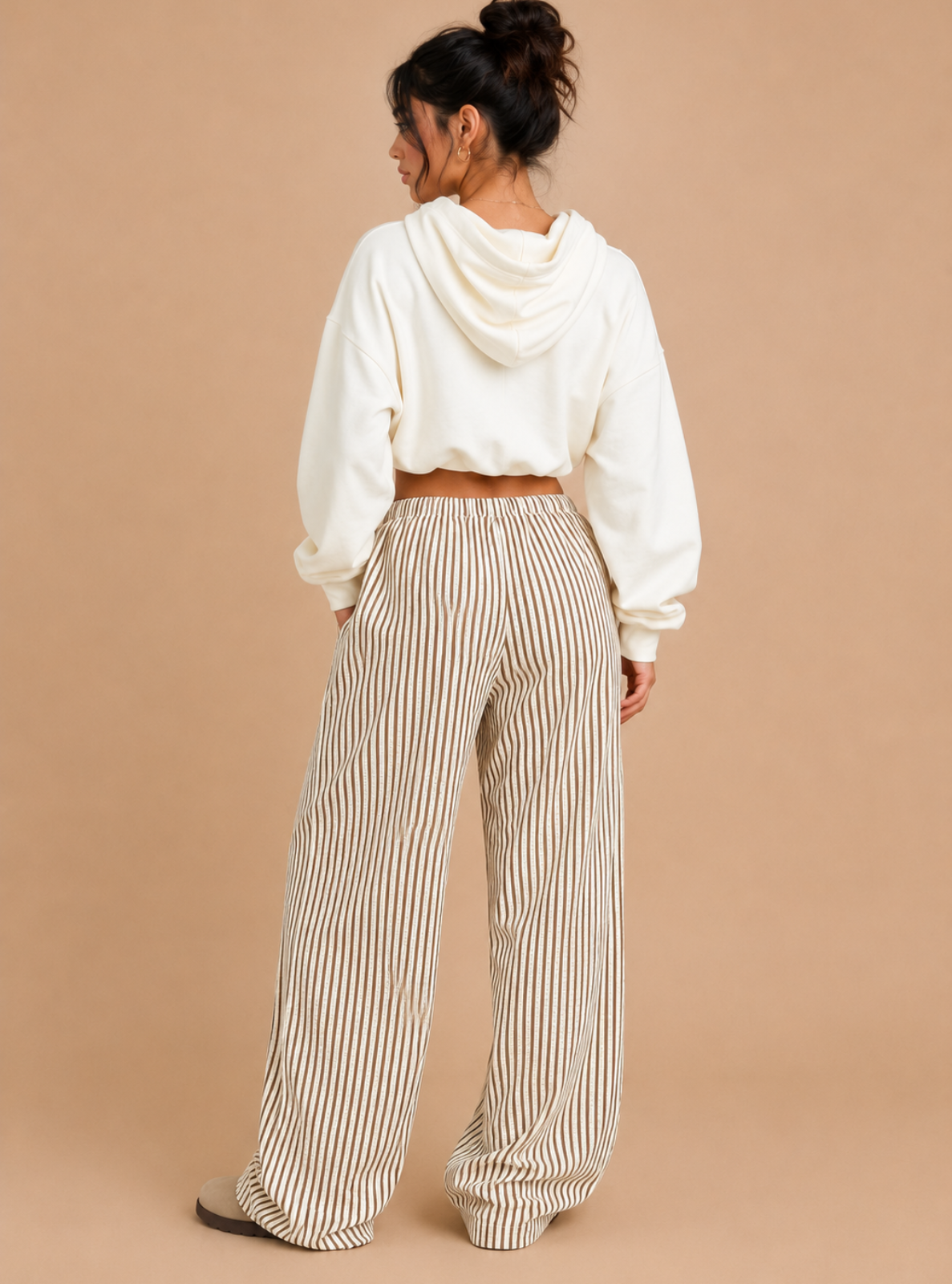 A woman shown from the back wearing a white cropped hoodie and beige-and-white vertically striped wide-leg pants, standing against a solid tan studio background.
