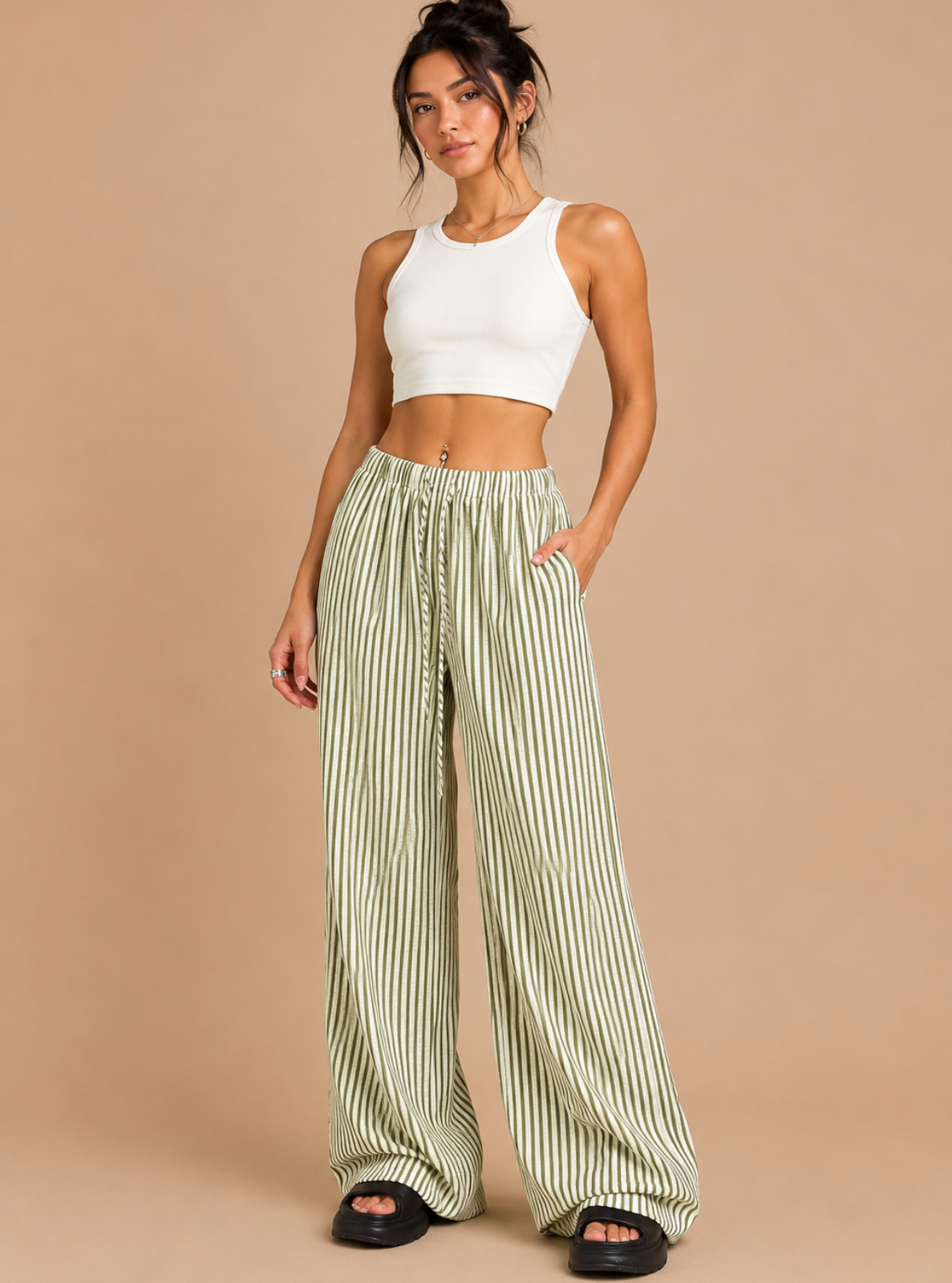 A woman modeling a white ribbed crop top and light green and white vertically striped wide-leg pants, paired with black slide sandals, against a solid beige studio background.