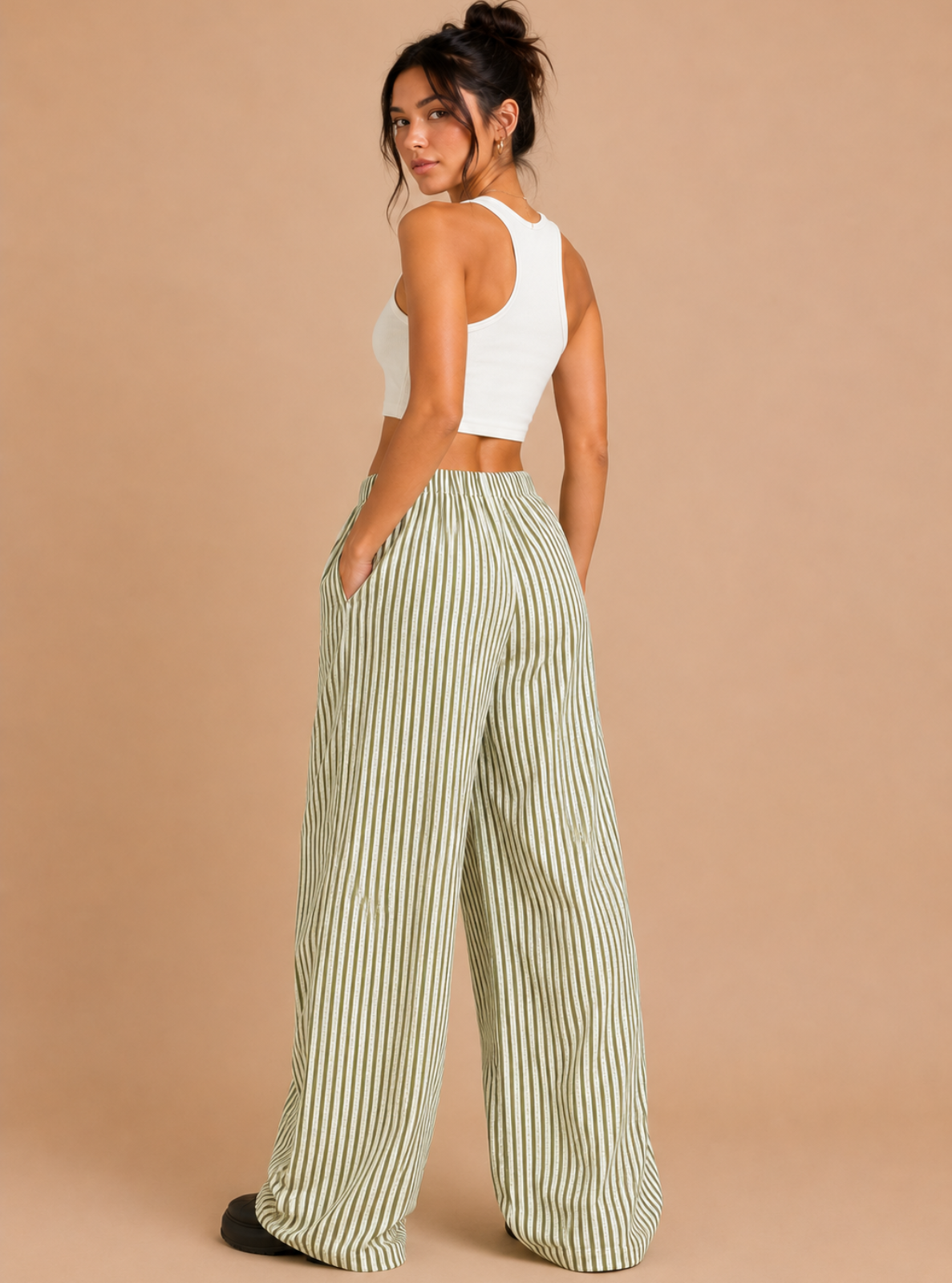A woman modeling a pair of wide-leg pants with vertical green and white stripes, paired with a white crop top, standing against a solid beige background.