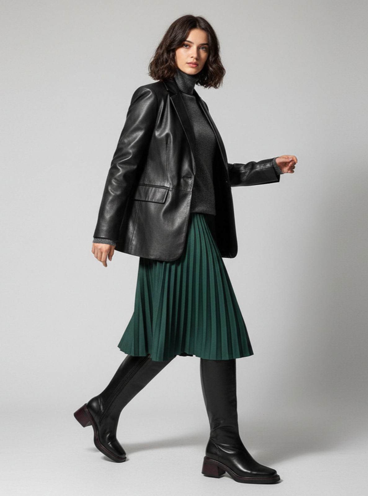 Woman wearing a black leather jacket, black turtleneck, green pleated skirt, and black boots on a gray background