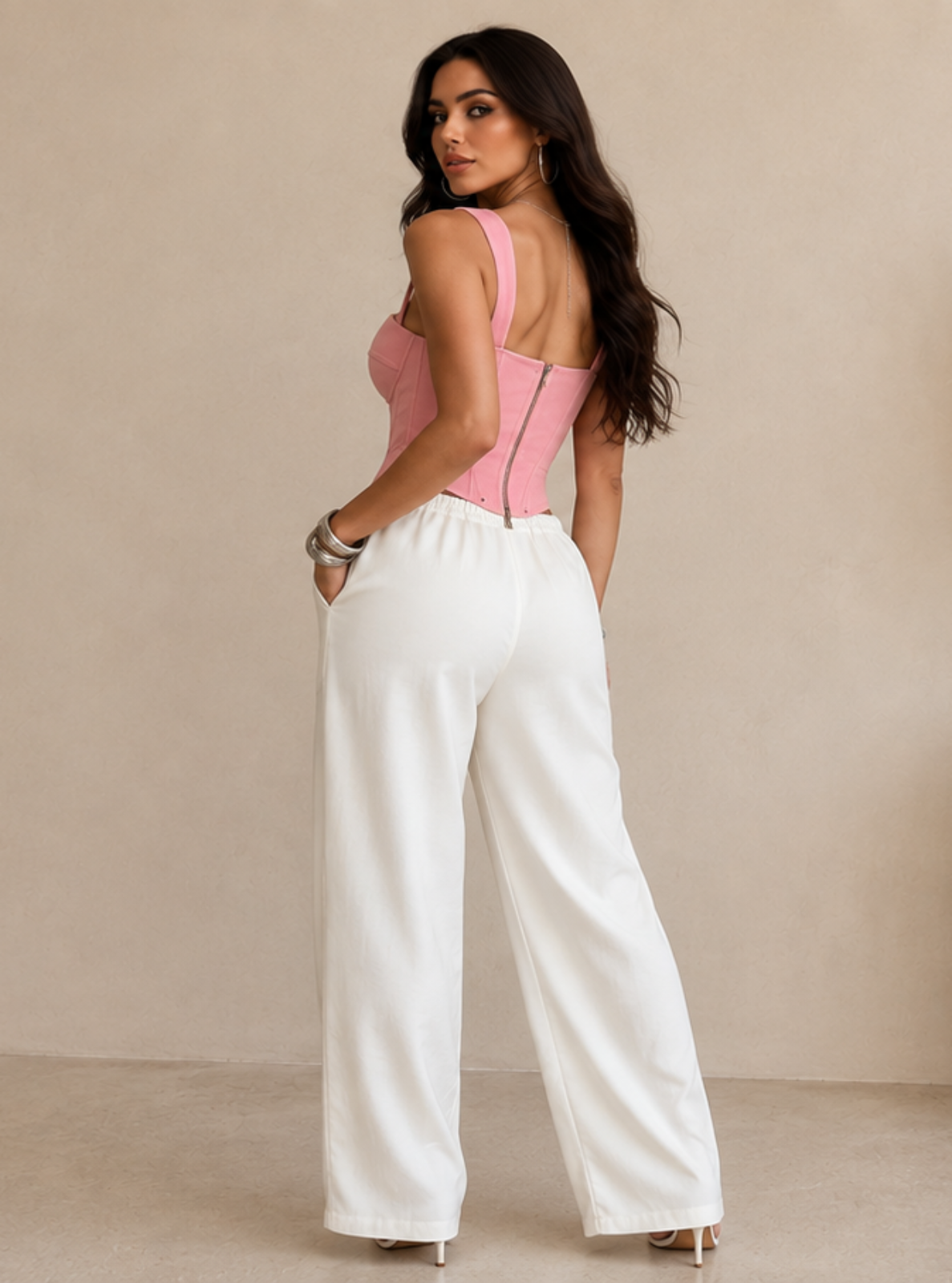 A woman stands in a minimalist beige room wearing a pink sleeveless backless top and white wide-leg pants, with a tall vase of dried grass beside her.