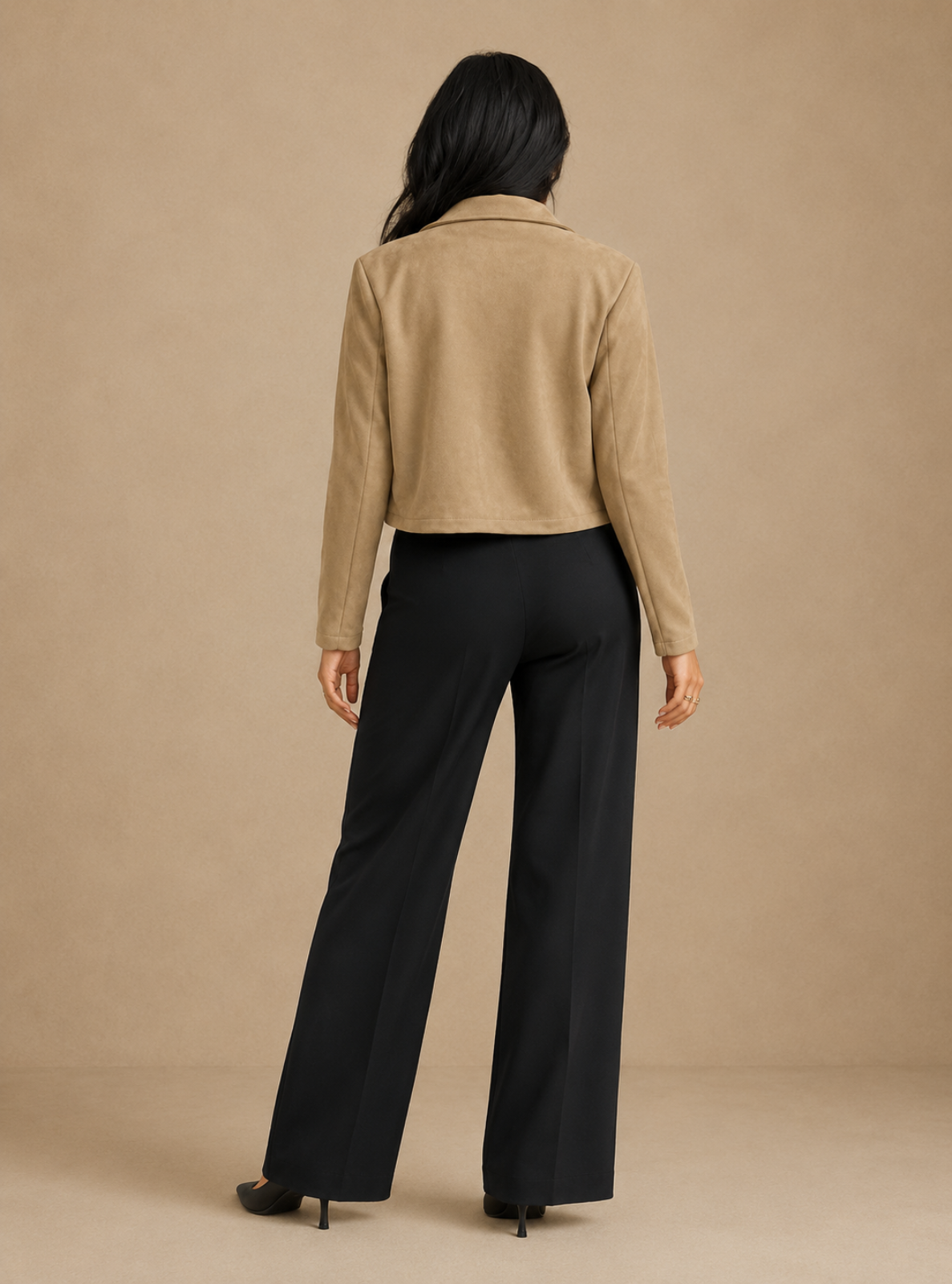 A woman shown from the back wearing a cropped tan suede jacket and black wide-leg trousers, standing against a plain beige studio backdrop.