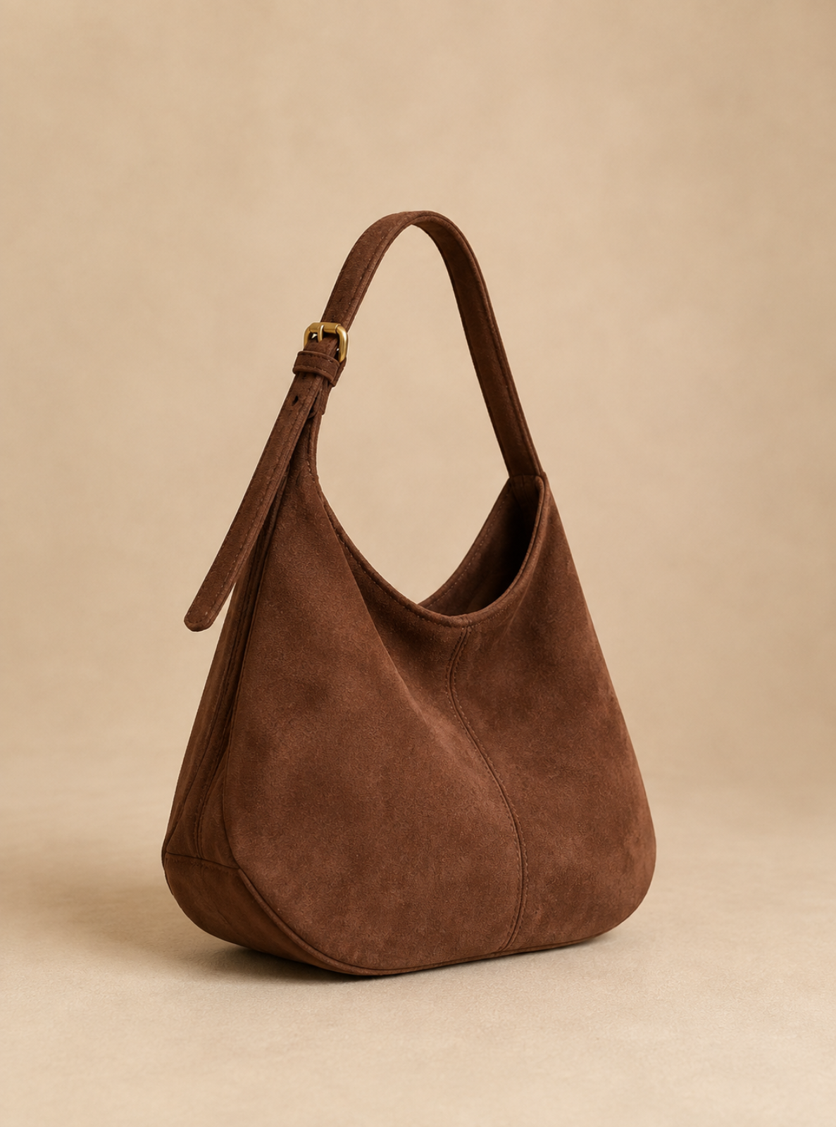 A brown suede hobo shoulder bag with a single adjustable strap and a small gold-tone buckle, shown on a warm beige studio background.
