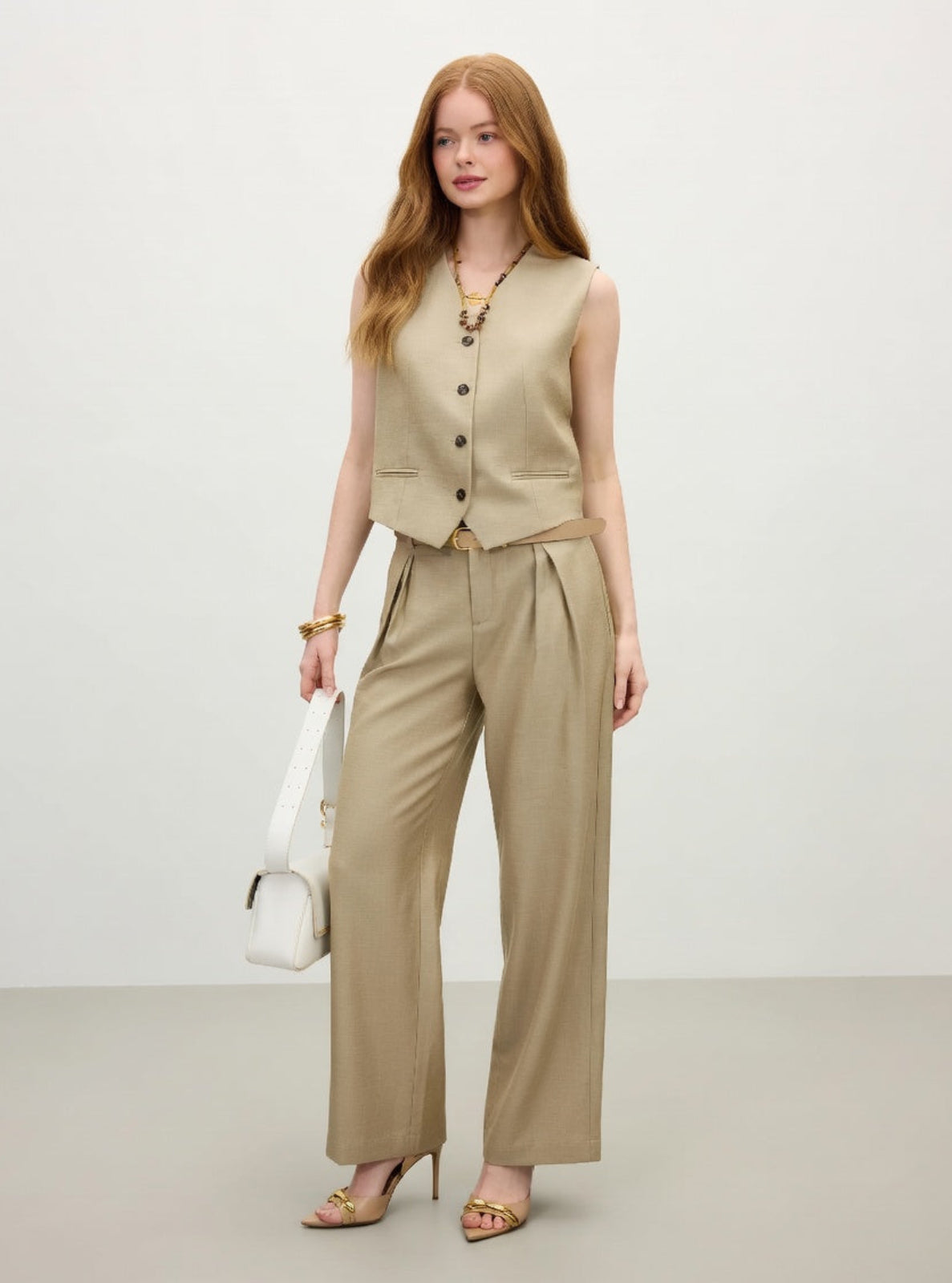 Woman wearing a beige vest and pants with a white handbag on a light background