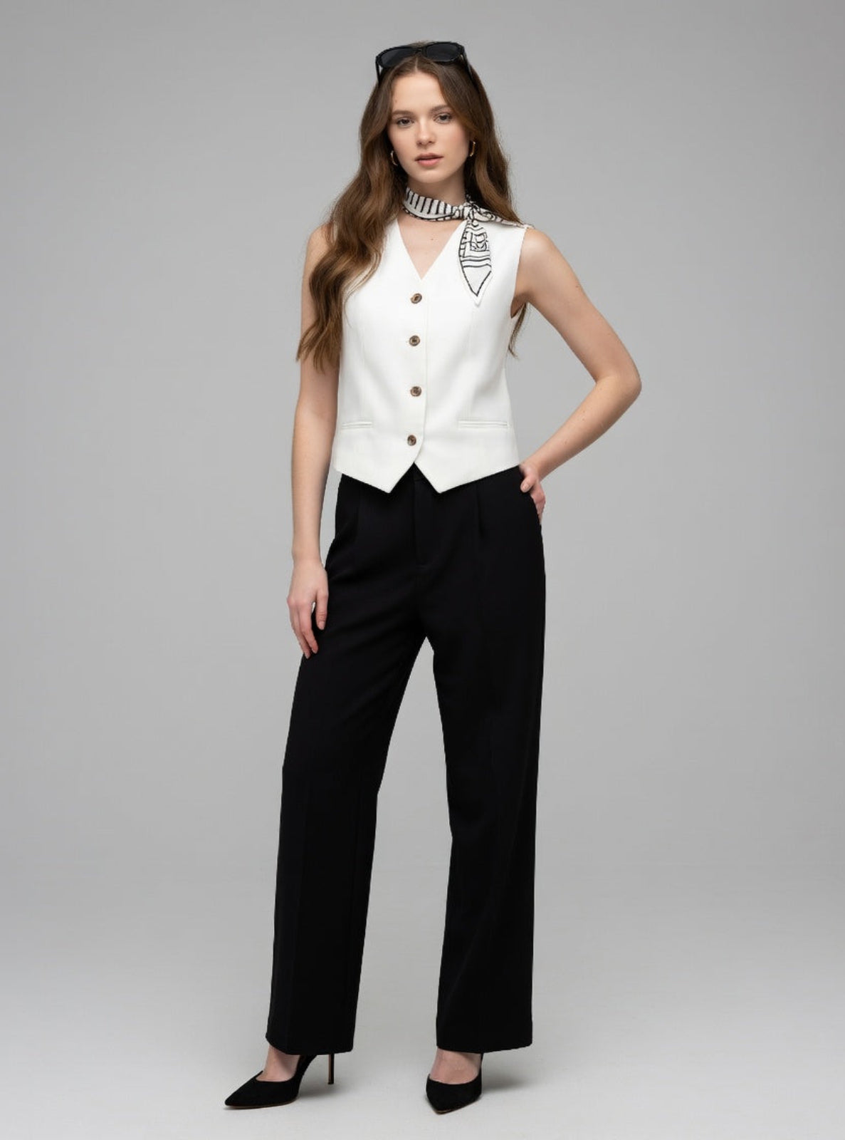 Woman wearing a white sleeveless top with buttons and black pants on a gray background