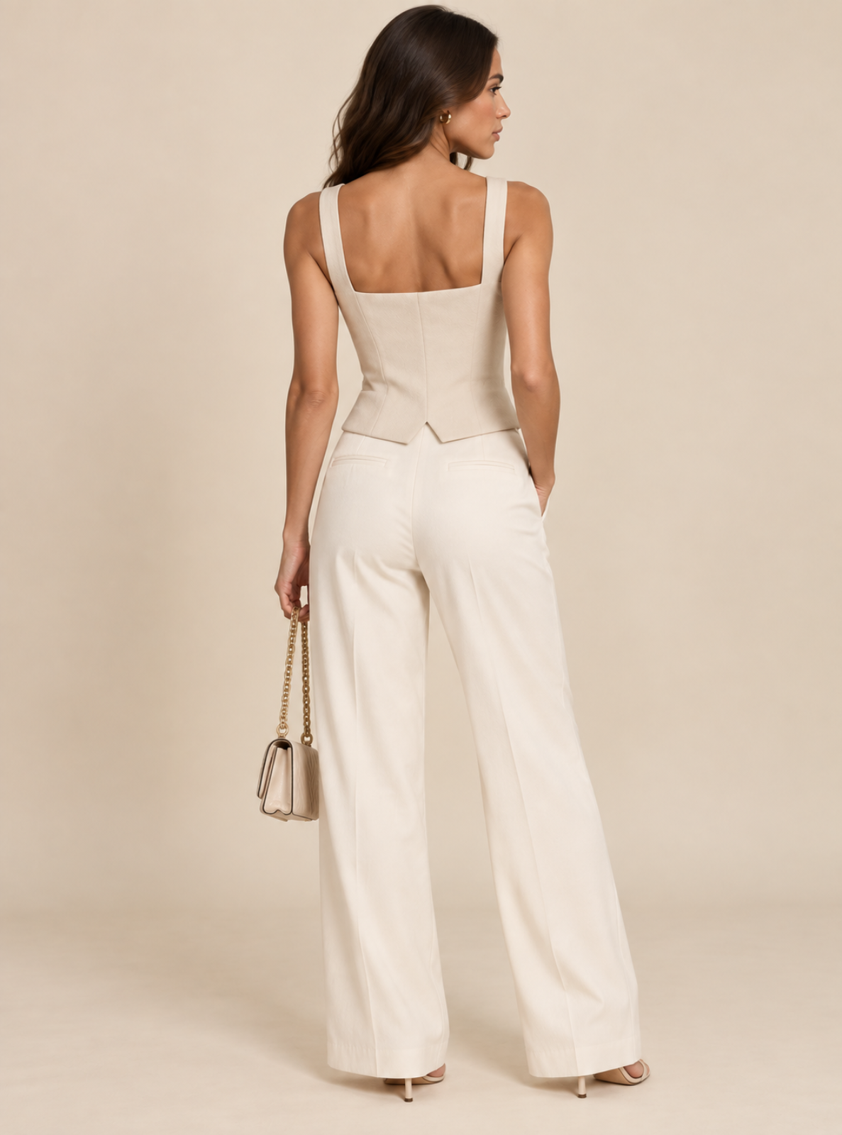 A woman shown from the back wearing a cream-colored sleeveless jumpsuit with a structured corset-style bodice and wide-leg pants, standing against a plain beige studio background and holding a small metallic handbag.