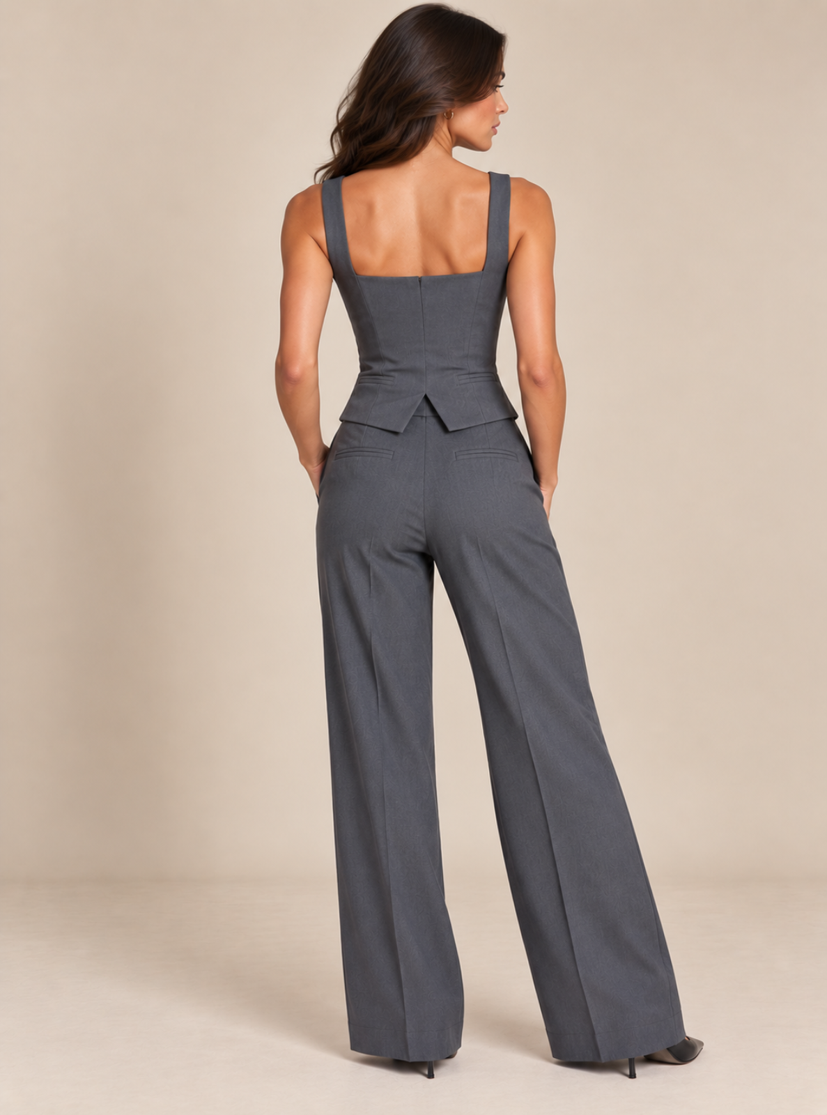 A woman shown from the back wearing a dark gray sleeveless wide-leg jumpsuit with a structured bodice and belt detail, set against a plain beige studio background.