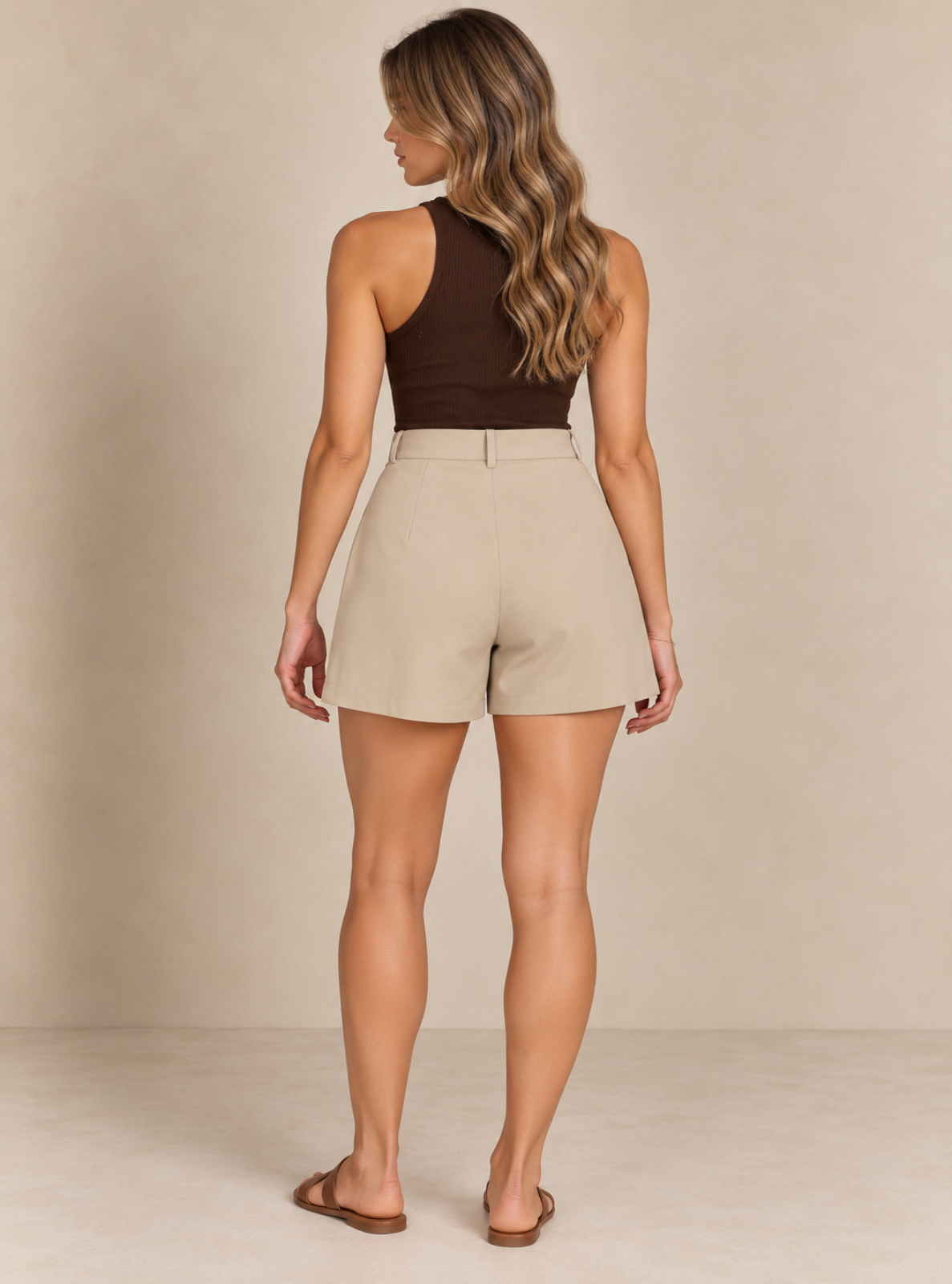 A woman shown from the back wearing a dark brown halter top and beige high-waisted shorts, standing against a neutral beige studio backdrop.