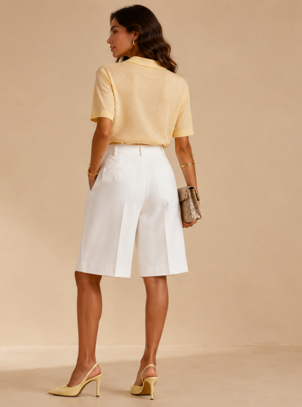 A woman stands with her back to the camera wearing a pale yellow short-sleeve top, white high-waisted shorts, and yellow heels, holding a small metallic clutch against a warm beige studio backdrop with soft leaf shadows.