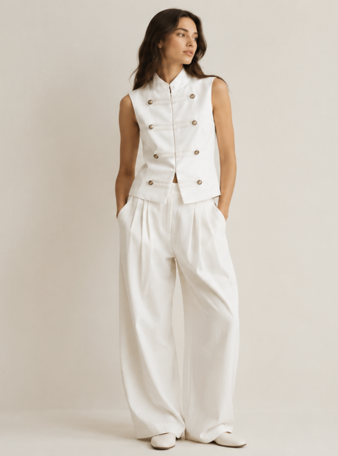 A woman stands against a plain light beige studio background wearing a white sleeveless double-breasted vest with dark buttons and matching wide-leg white trousers.
