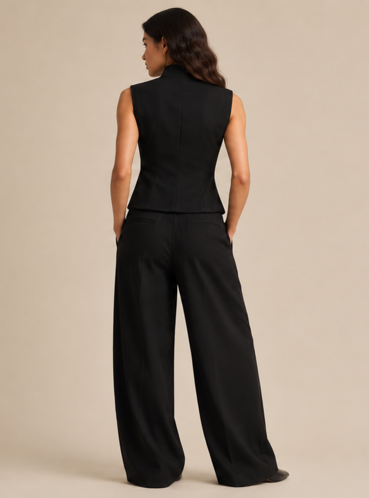 A woman viewed from the back wearing a black sleeveless top and matching wide-leg pants against a plain beige studio background.