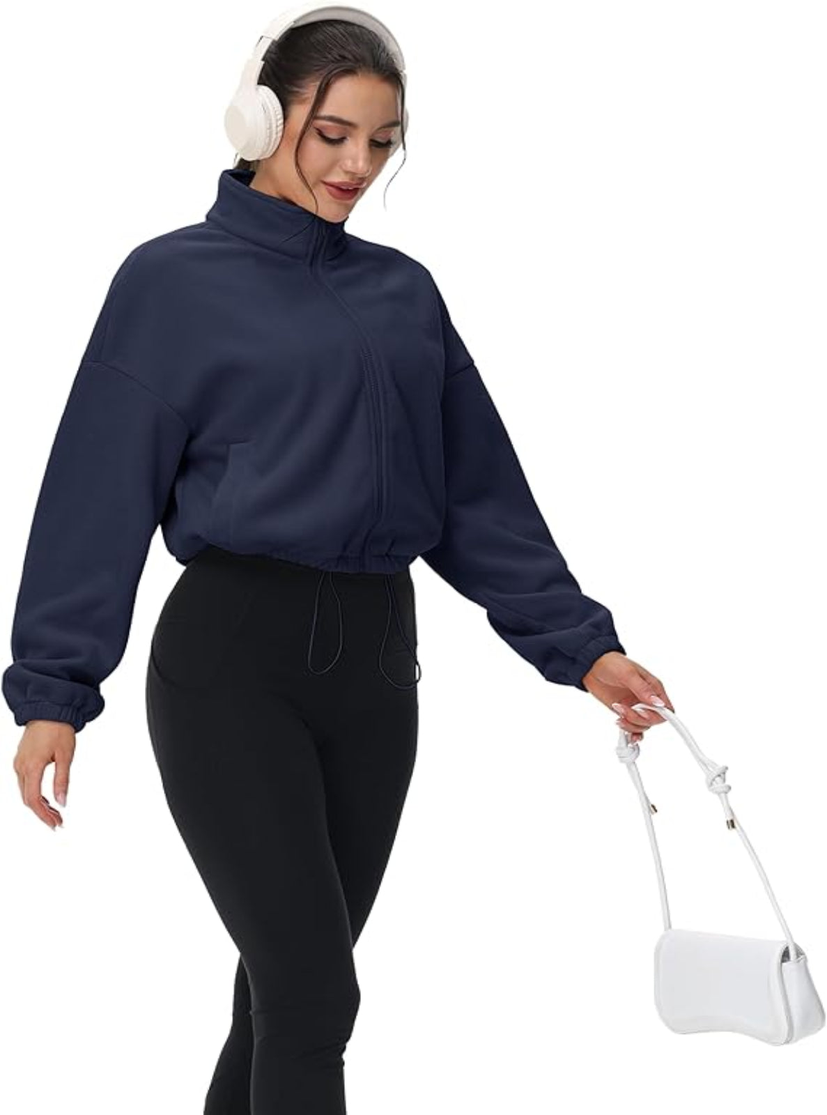 Woman wearing a navy blue long-sleeve top and black pants, holding a white handbag on a white background.