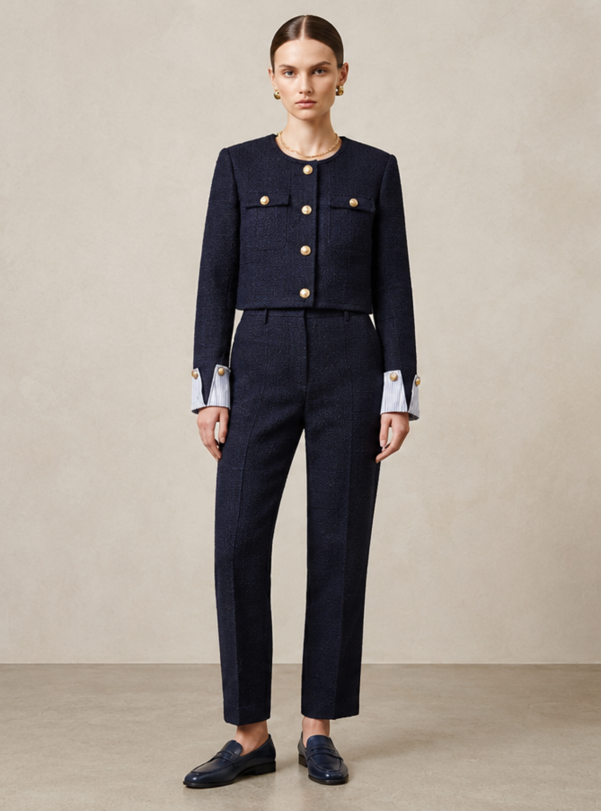 A model stands in a minimalist studio wearing a dark navy blue two-piece suit with a cropped jacket and matching trousers. The jacket features light blue and white striped button accents at the collar and cuffs.