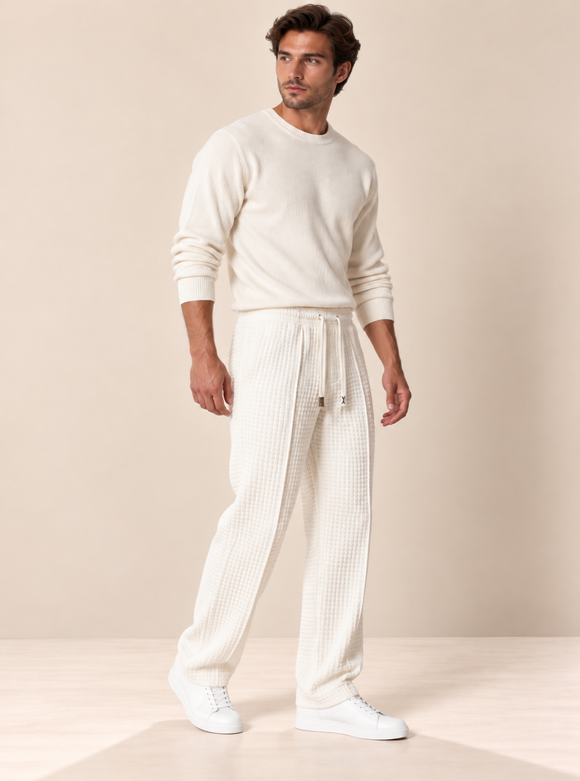 A man modeling a cream-colored knit crewneck sweater and matching textured waffle-knit drawstring sweatpants, paired with white sneakers, against a warm beige studio background.