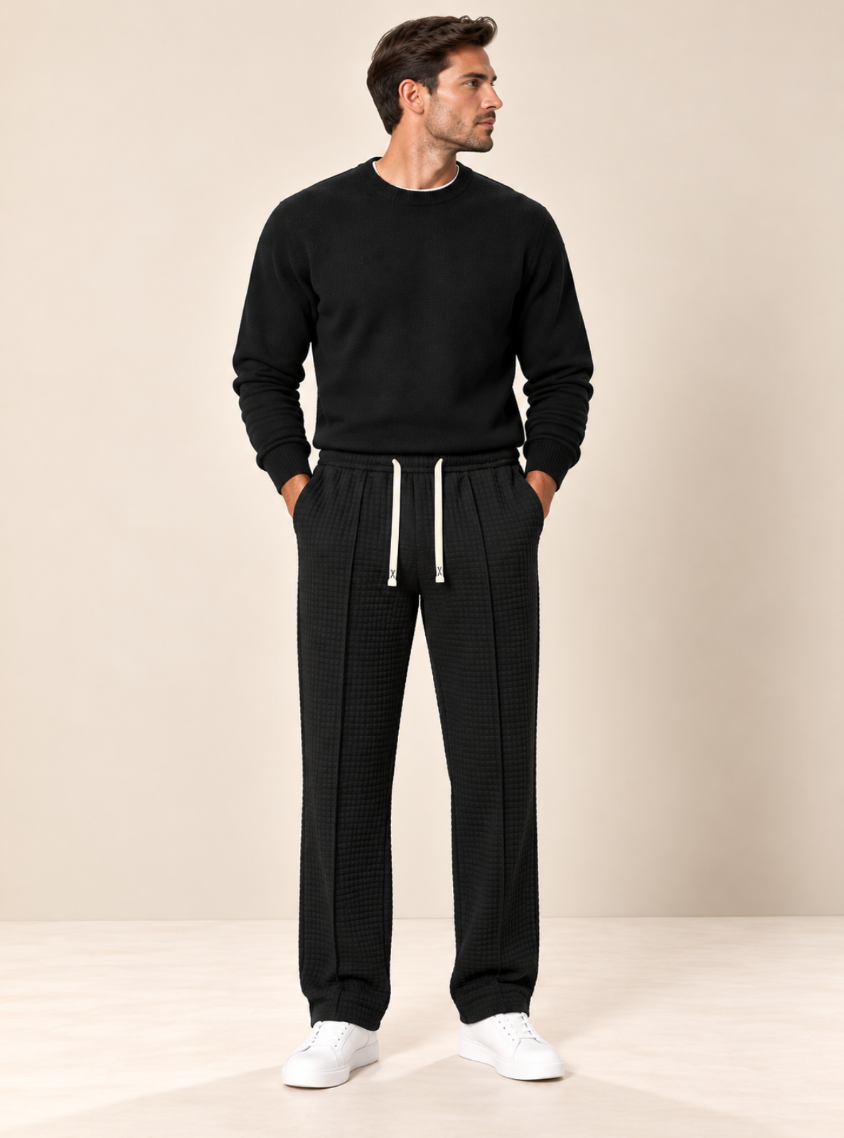 A man modeling a black crewneck sweatshirt and textured black drawstring sweatpants with white drawstrings, paired with white sneakers, against a neutral beige studio background.