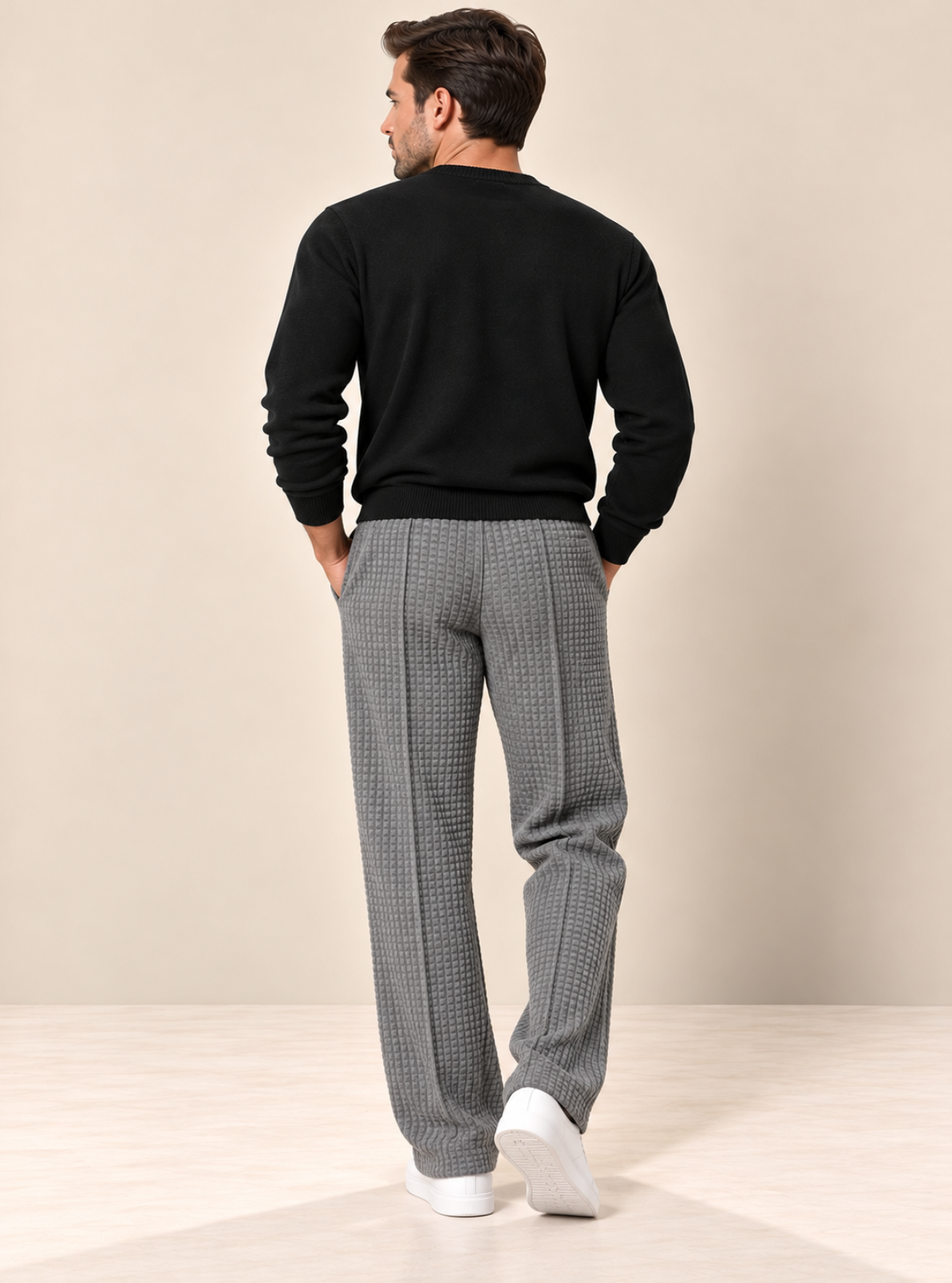 A man viewed from behind wearing a black long-sleeve crewneck sweater and gray houndstooth trousers, paired with white sneakers, against a neutral beige studio background.