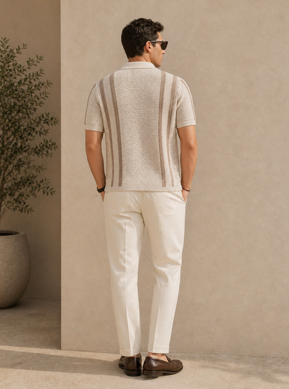 Man wearing a striped shirt and white pants standing against a beige wall.