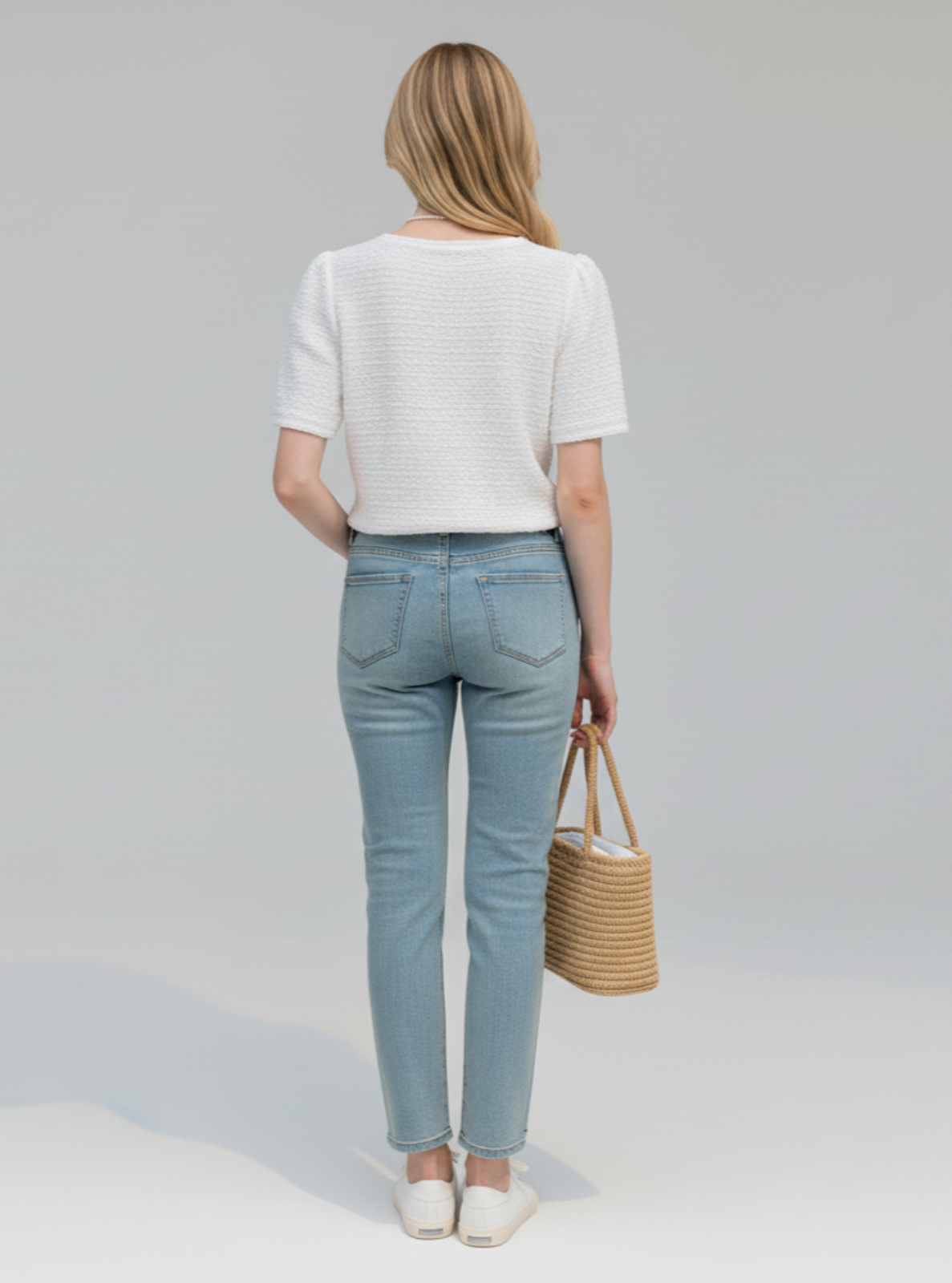 Woman wearing light blue jeans and a white top, holding a woven bag on a plain background
