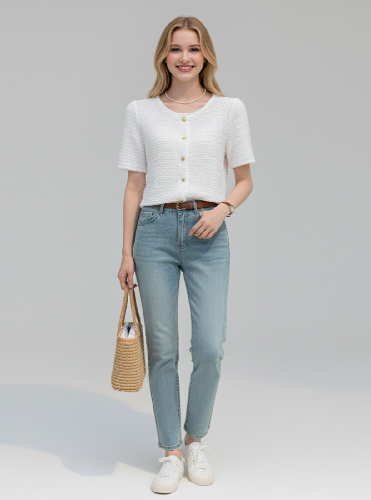 Woman wearing a white top and light blue jeans, holding a woven bag on a plain background