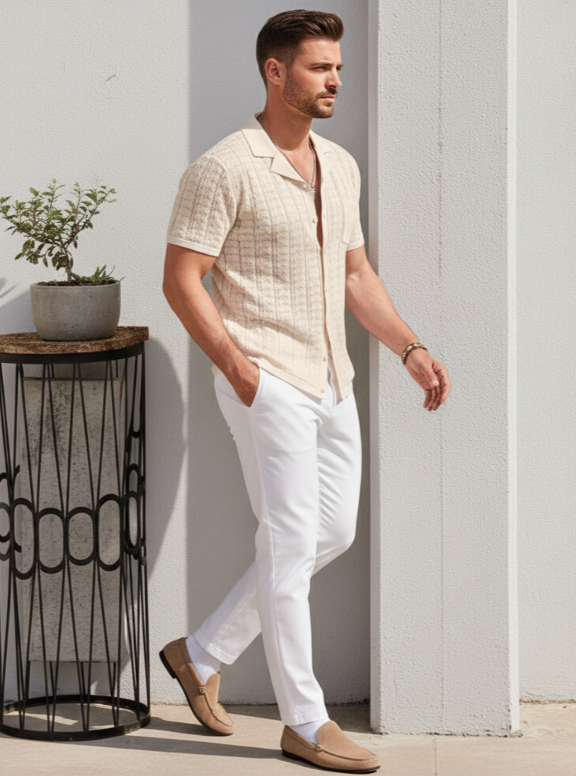 Man in beige shirt and white pants standing next to a small plant stand against a light-colored wall.