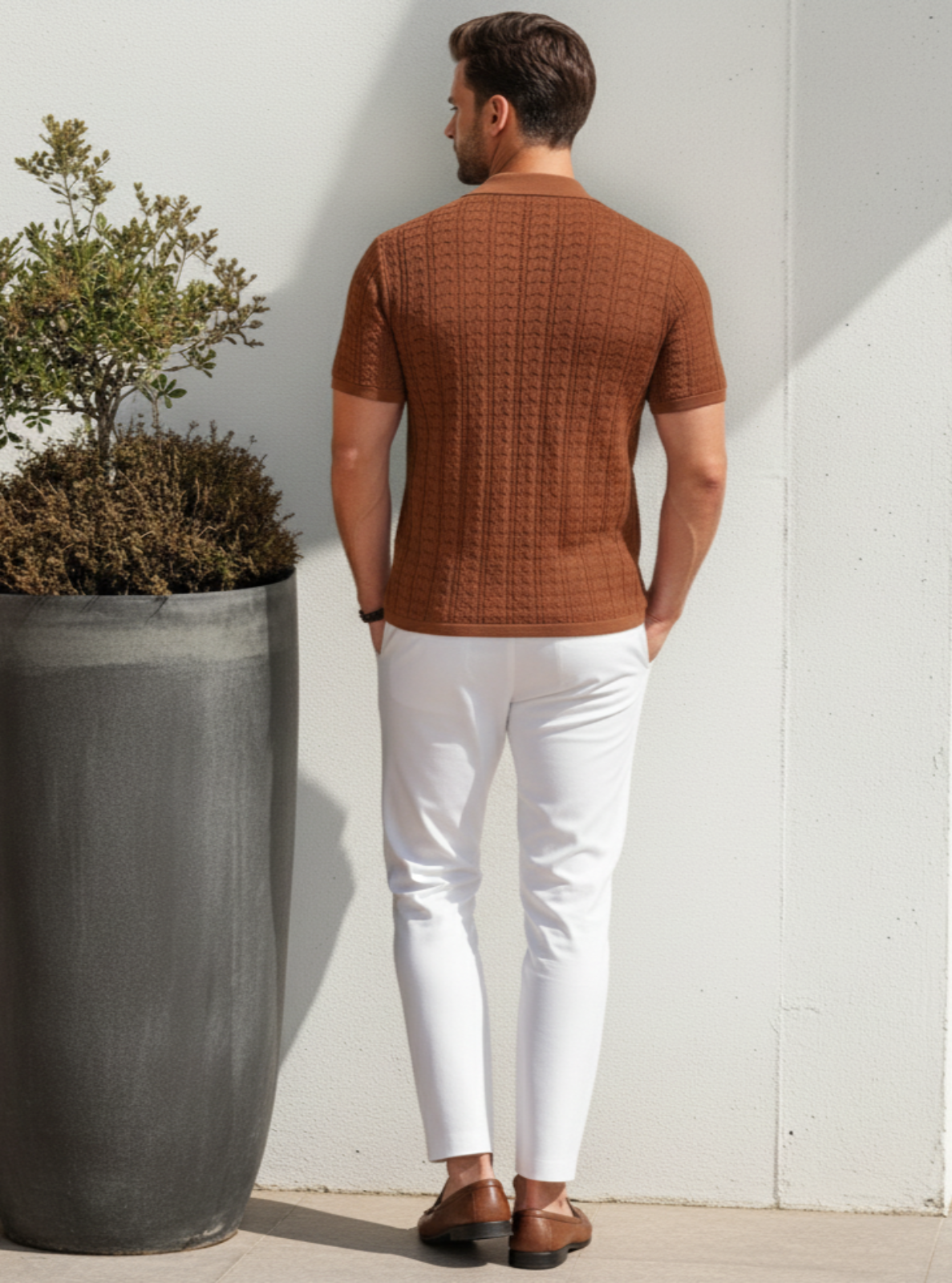 Man in a brown textured shirt and white pants standing next to a large planter against a white wall.