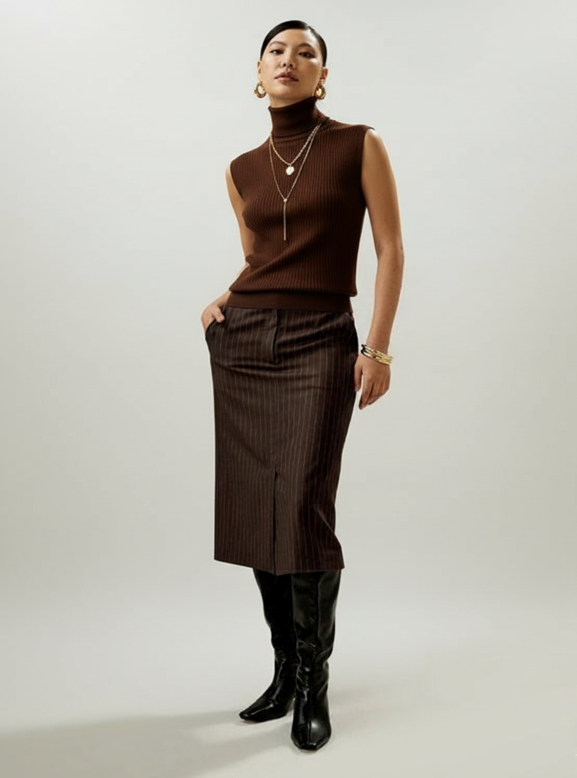 Woman wearing a brown sleeveless top and pinstripe skirt with black knee-high boots on a plain background