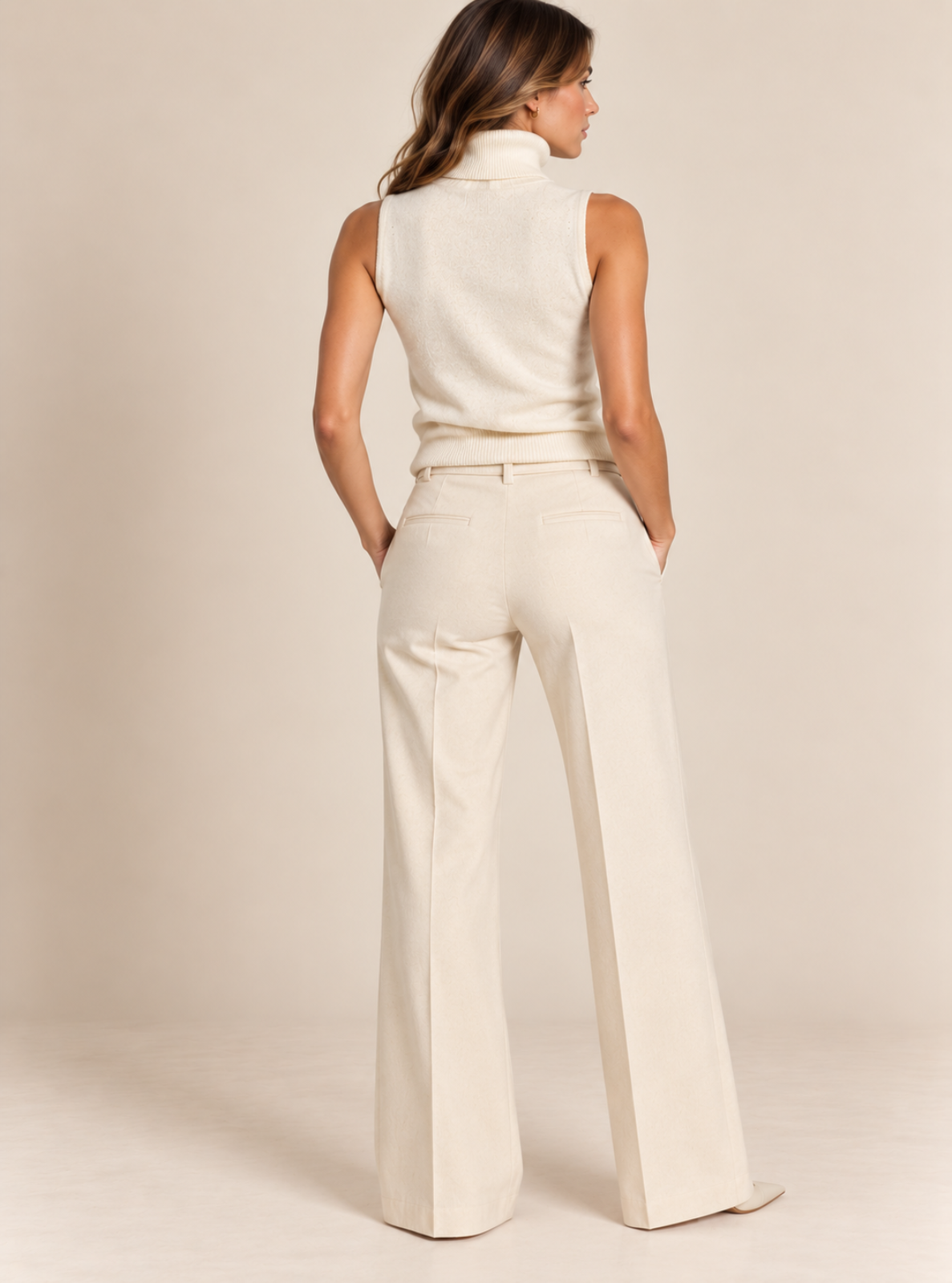 A woman shown from the back wearing a cream-colored sleeveless mock-neck top and matching high-waisted, wide-leg trousers against a solid beige studio background.