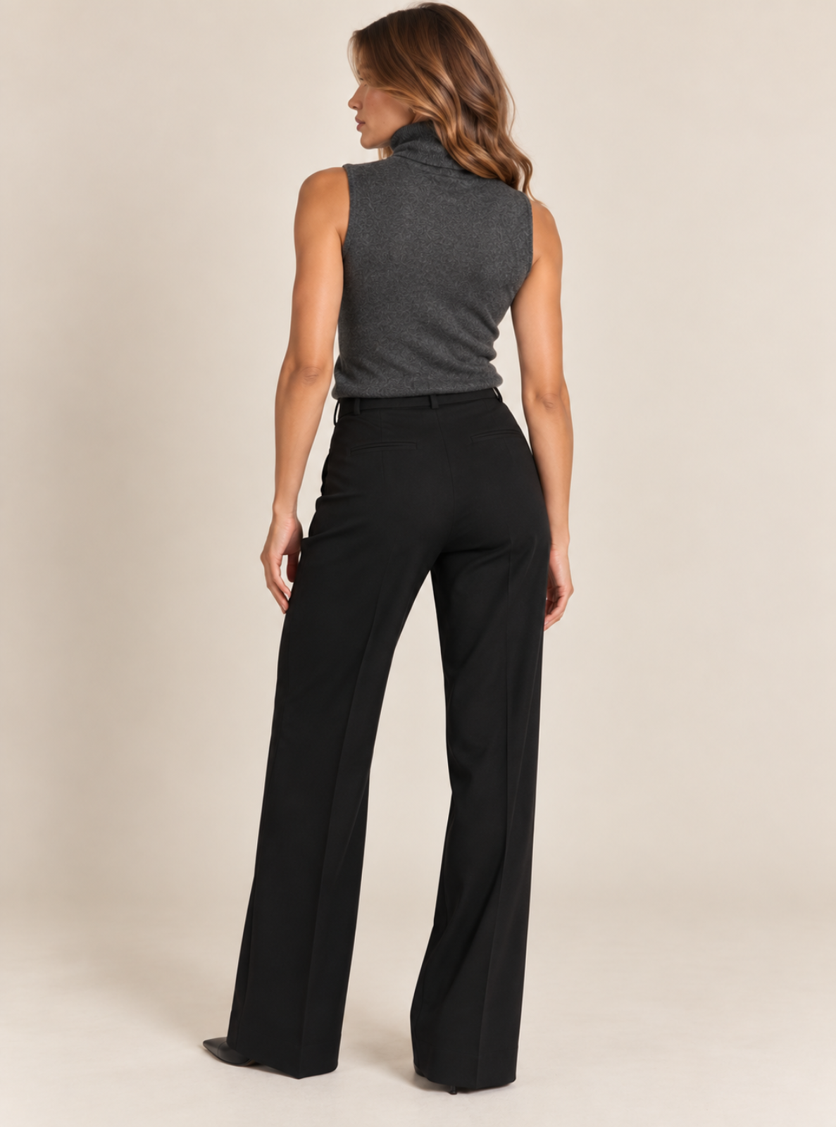 A woman shown from the back wearing a sleeveless dark gray mock-neck top and black wide-leg trousers, standing against a plain beige studio background.