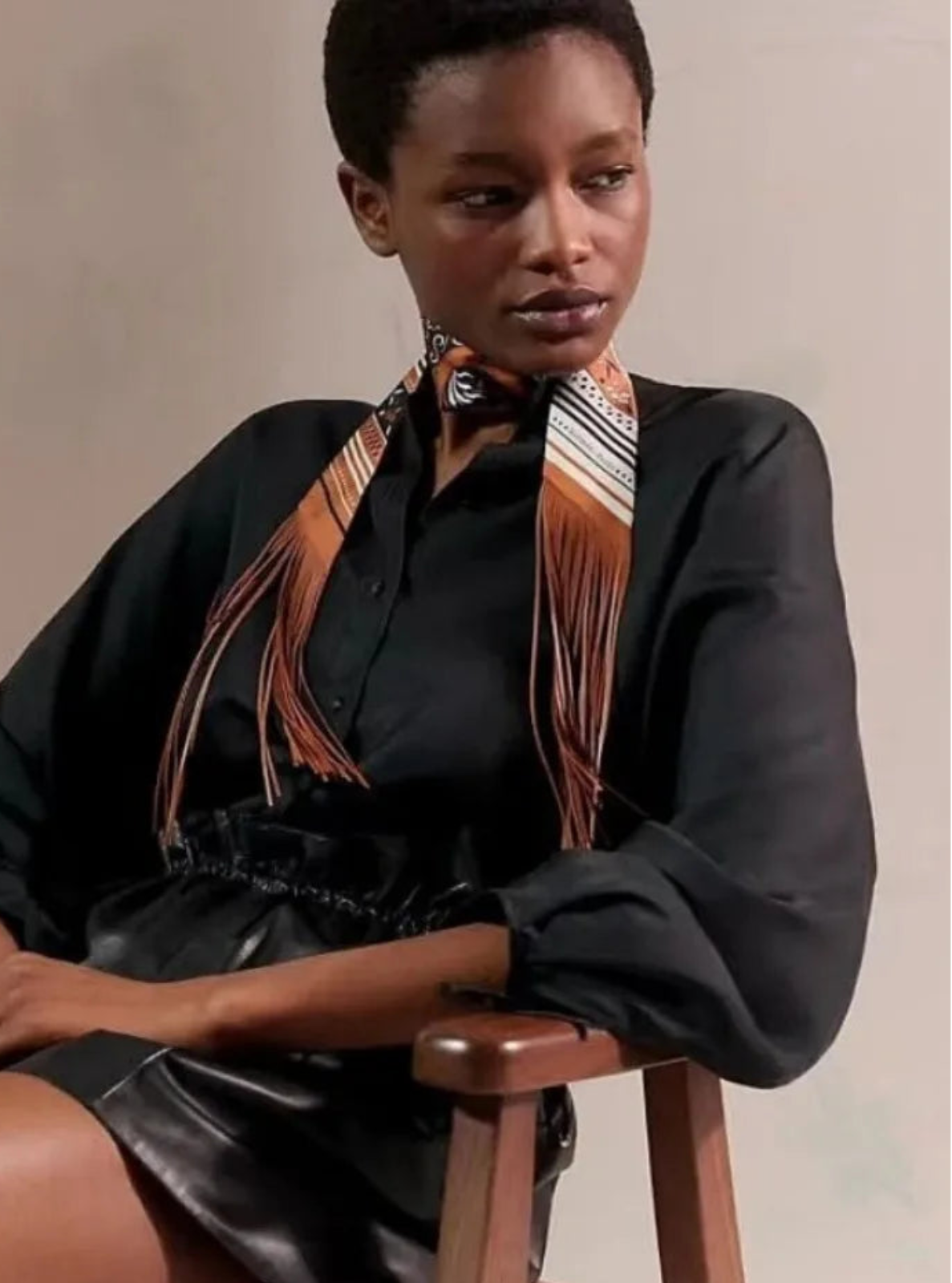 Woman sitting on a chair wearing a black outfit with a patterned scarf, against a neutral background