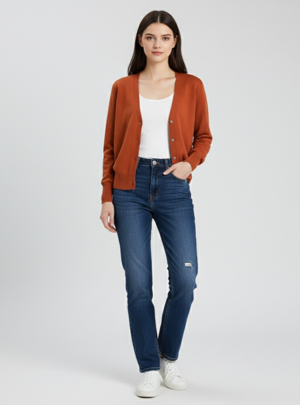 Woman wearing a rust-colored cardigan, white top, blue jeans, and white sneakers on a light gray background