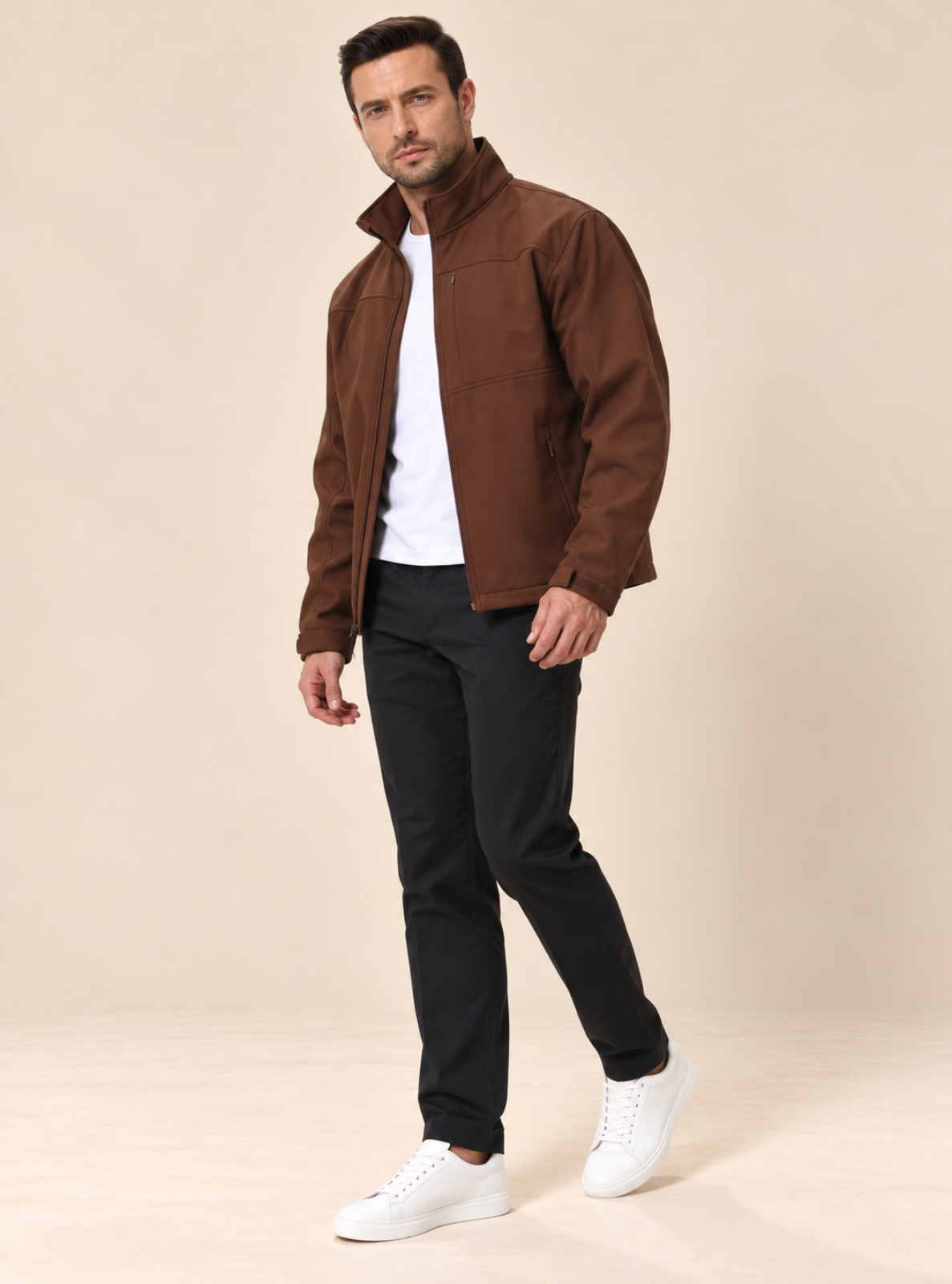 A man modeling a brown zip-up utility jacket over a white t-shirt, black trousers, and white sneakers, standing against a solid beige studio background.