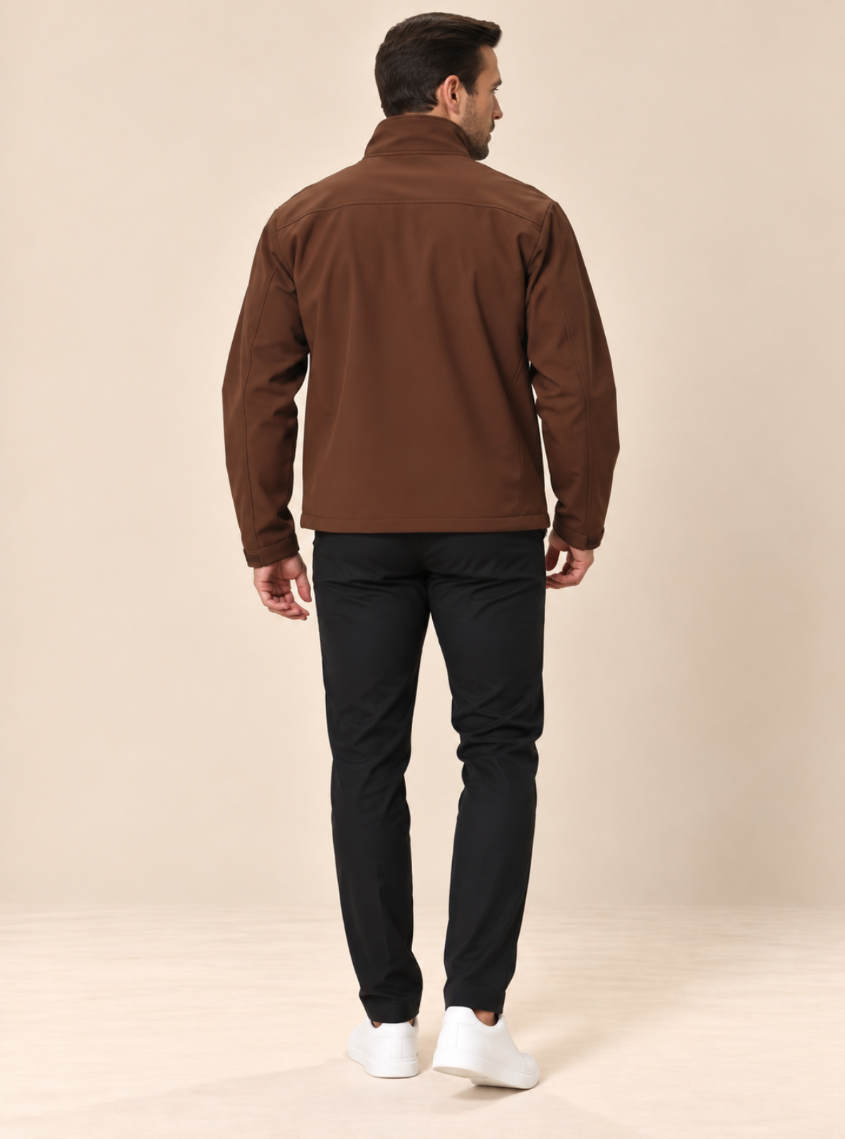 A man shown from the back wearing a brown zip-up jacket, black pants, and white sneakers against a plain beige studio background.