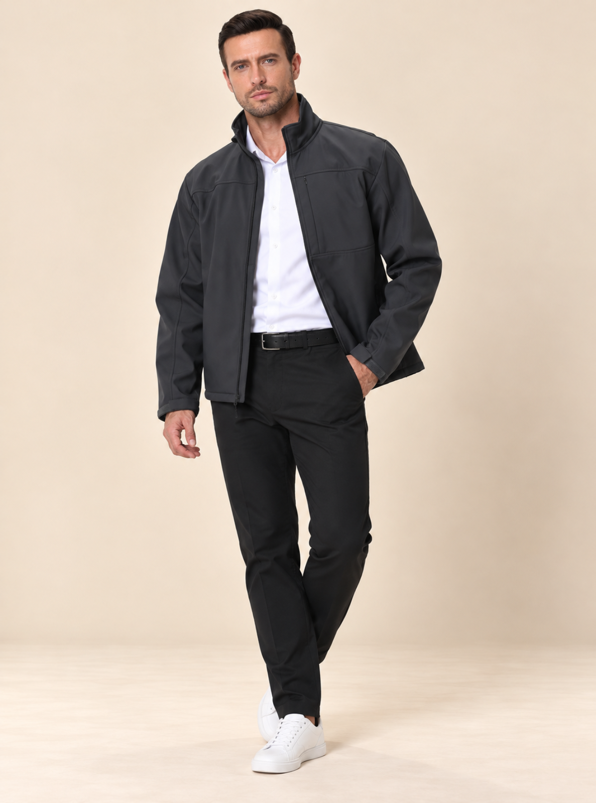 A man modeling a dark gray lightweight jacket over a white collared shirt, paired with black trousers and white sneakers, standing against a solid beige studio background.