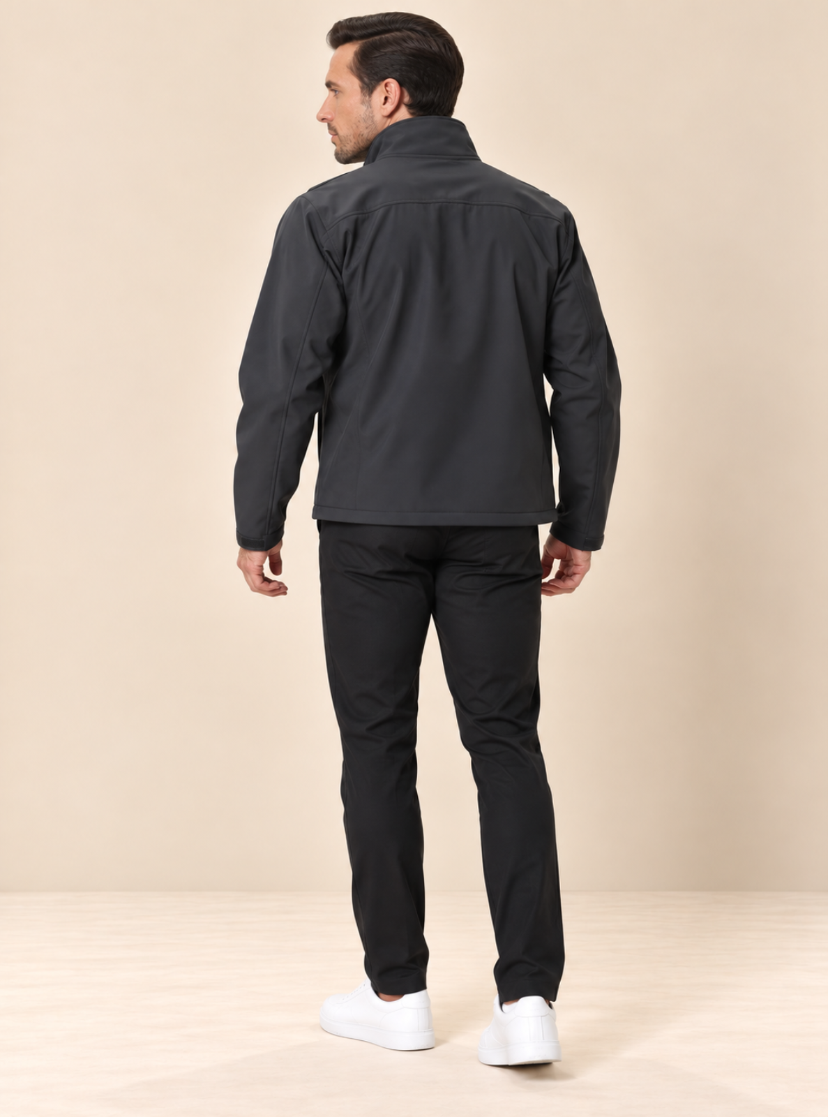 A man shown from the back wearing a dark gray zip-up jacket, black pants, and white sneakers, standing against a plain beige studio background.