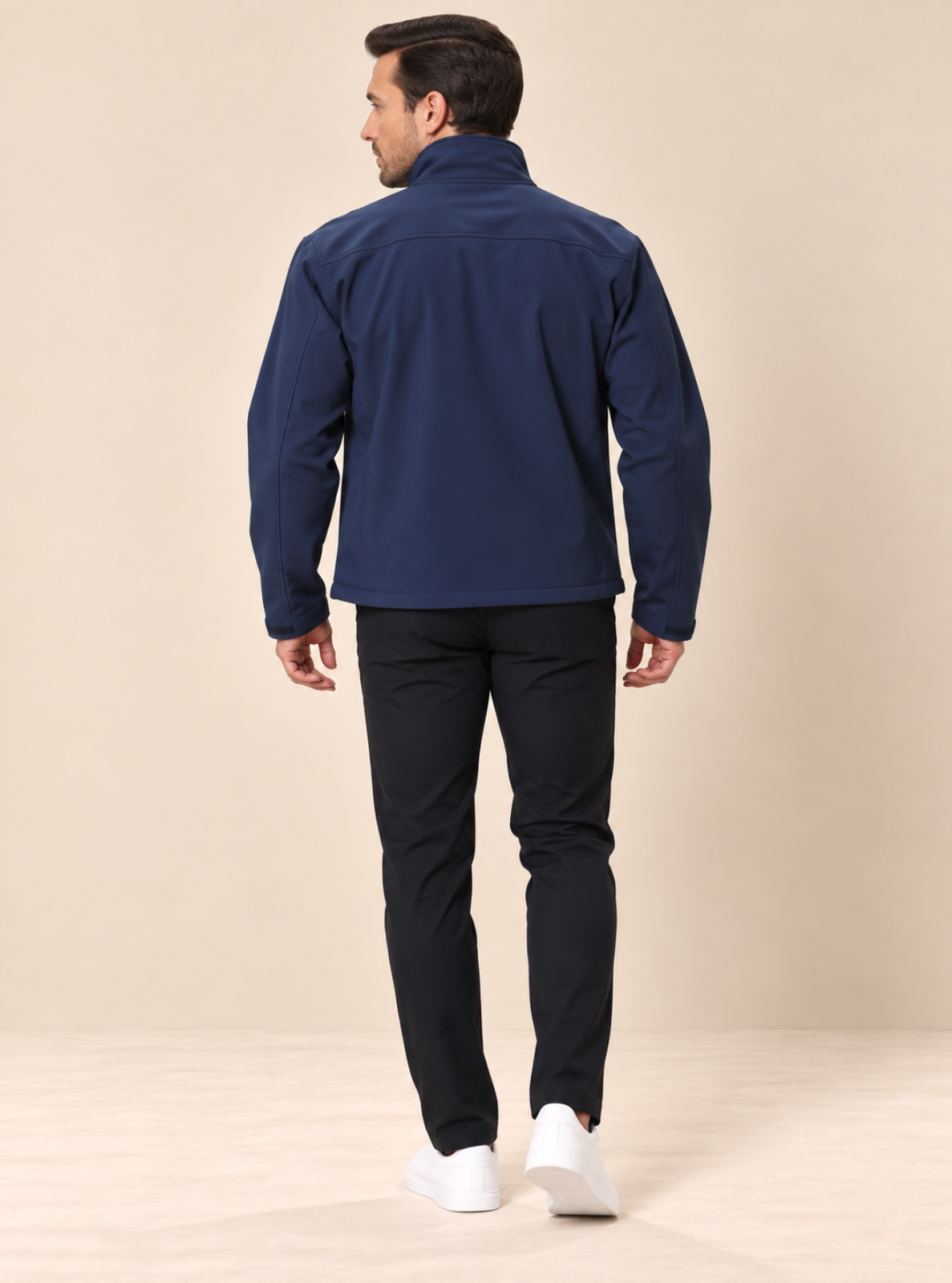 A man viewed from behind wearing a navy blue zip-up jacket, dark trousers, and white sneakers, standing against a plain beige studio background.