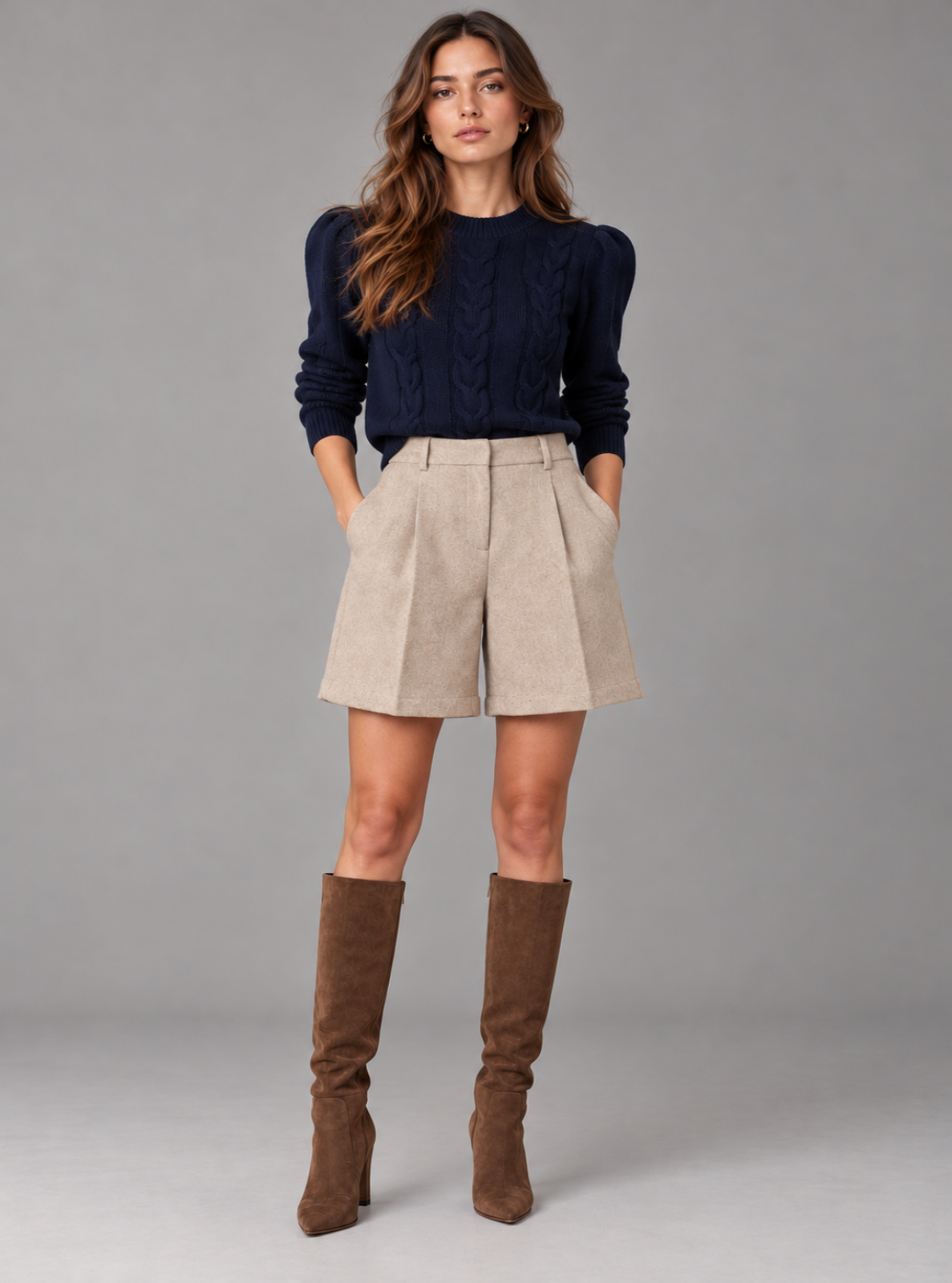 A woman modeling a navy blue cable-knit sweater, high-waisted beige pleated shorts, and brown knee-high suede boots against a solid gray studio background.