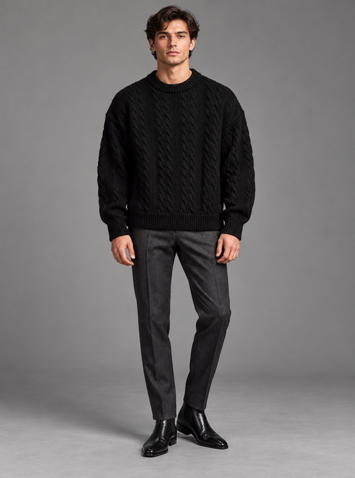 A man modeling a black cable-knit crewneck sweater, paired with dark gray trousers and black leather shoes, against a solid gray studio background.