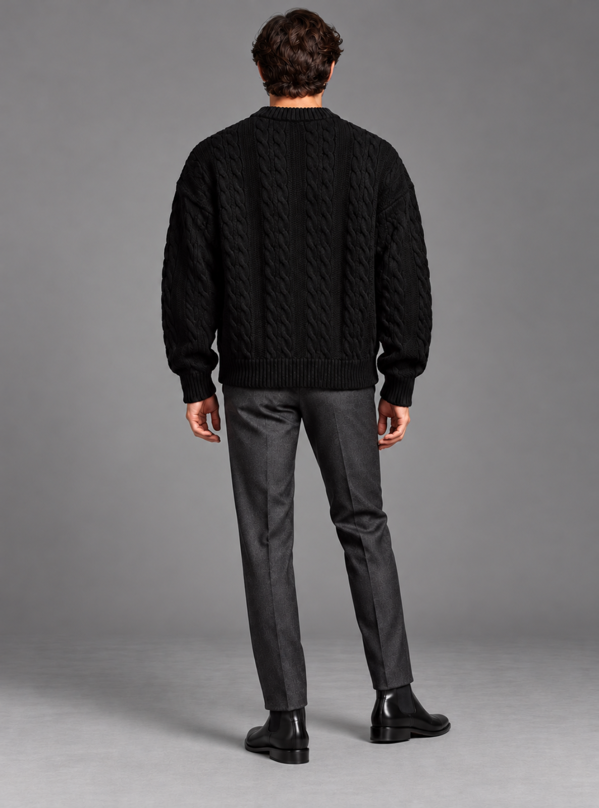 A man viewed from the back wearing a black cable-knit sweater, dark charcoal gray trousers, and black leather boots against a solid gray studio background.