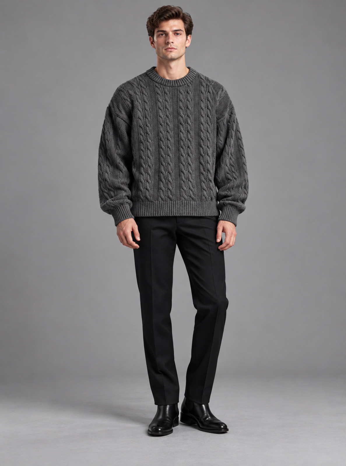 A man modeling a dark gray cable-knit crewneck sweater, paired with black trousers and black leather shoes, against a solid gray studio background.
